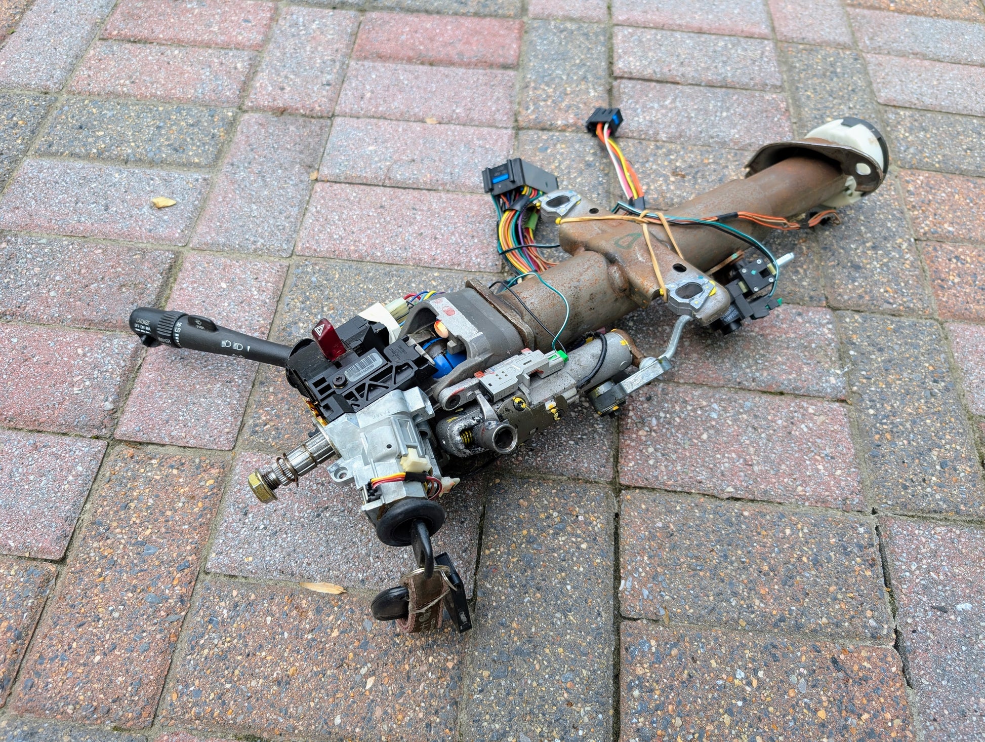 A General Motors OEM Steering Column w/KEY for 2000-2002 GM Full Size Trucks is lying on a brick-paved surface, complete with attached wiring and switches.