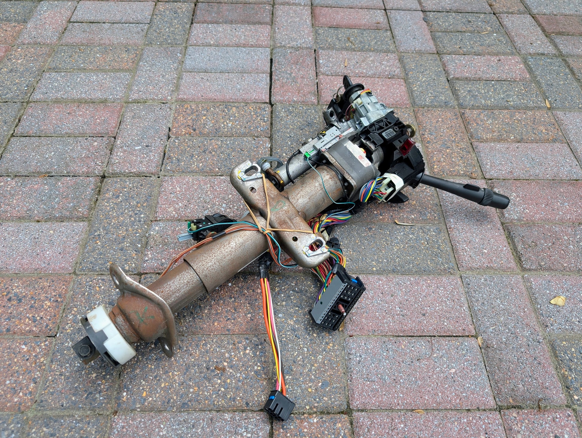 A rusty General Motors OEM Steering Column w/KEY for 2000-2002 GM Full Size Trucks, with exposed wires and connectors, rests on a patterned brick pavement.