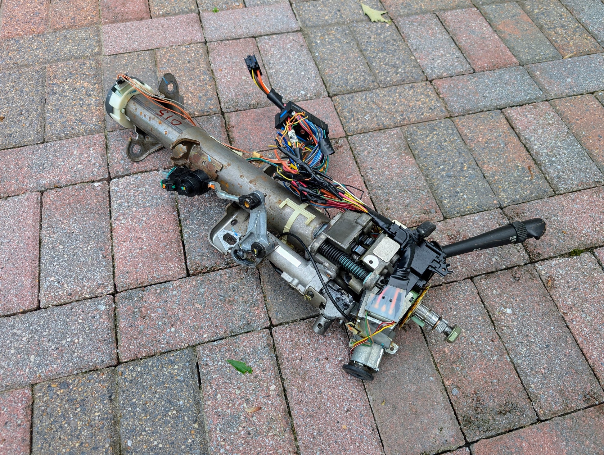A used General Motors OEM Steering Column for 2000-2002 GM Full Size Trucks with visible wiring, cables, and connectors is shown resting on a brick pavement.