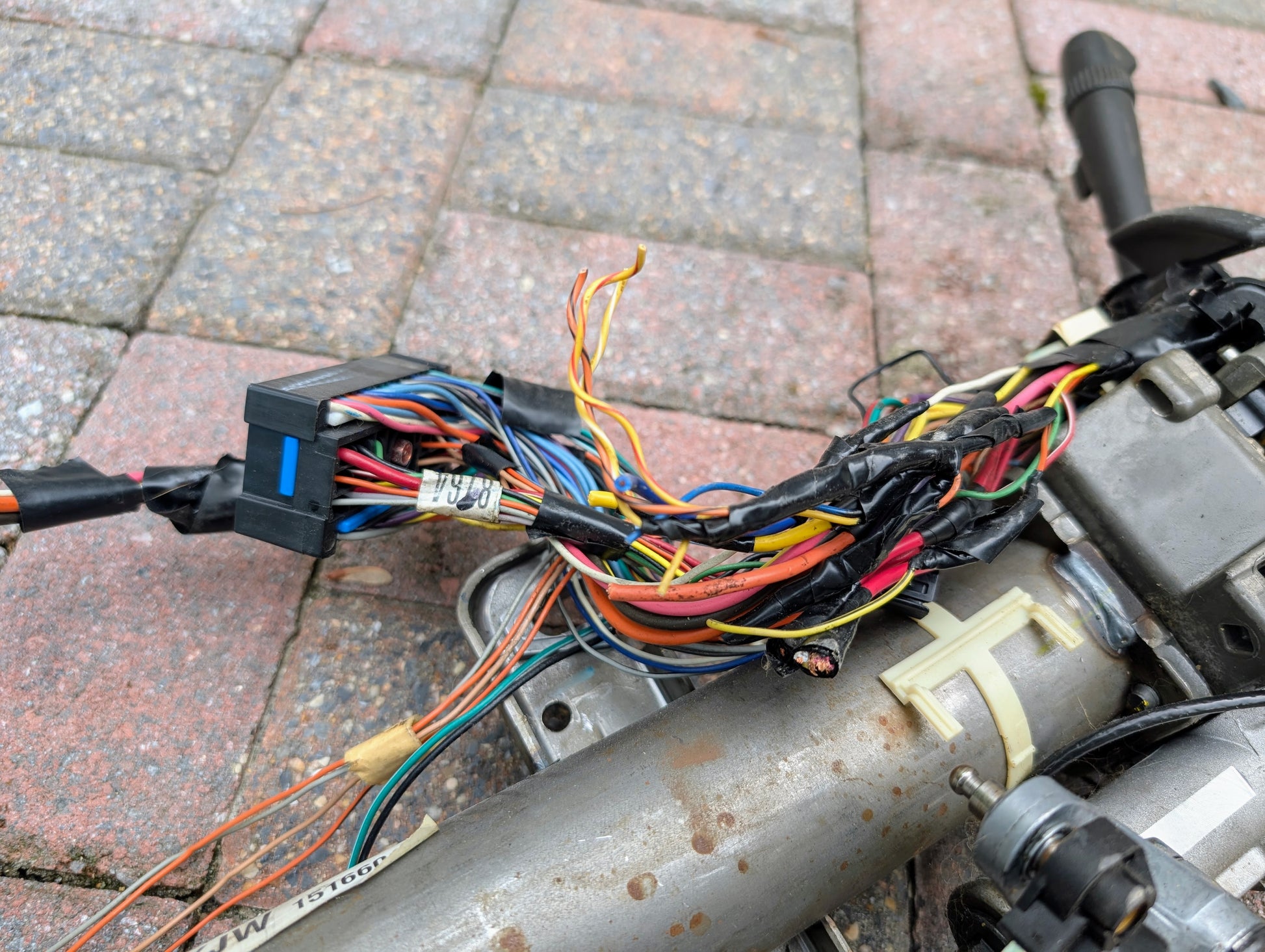 A close-up of multicolored wires, some wrapped in black tape, connected to a General Motors OEM Steering Column for 2000-2002 GM Full Size Trucks, resting on a brick-patterned surface.