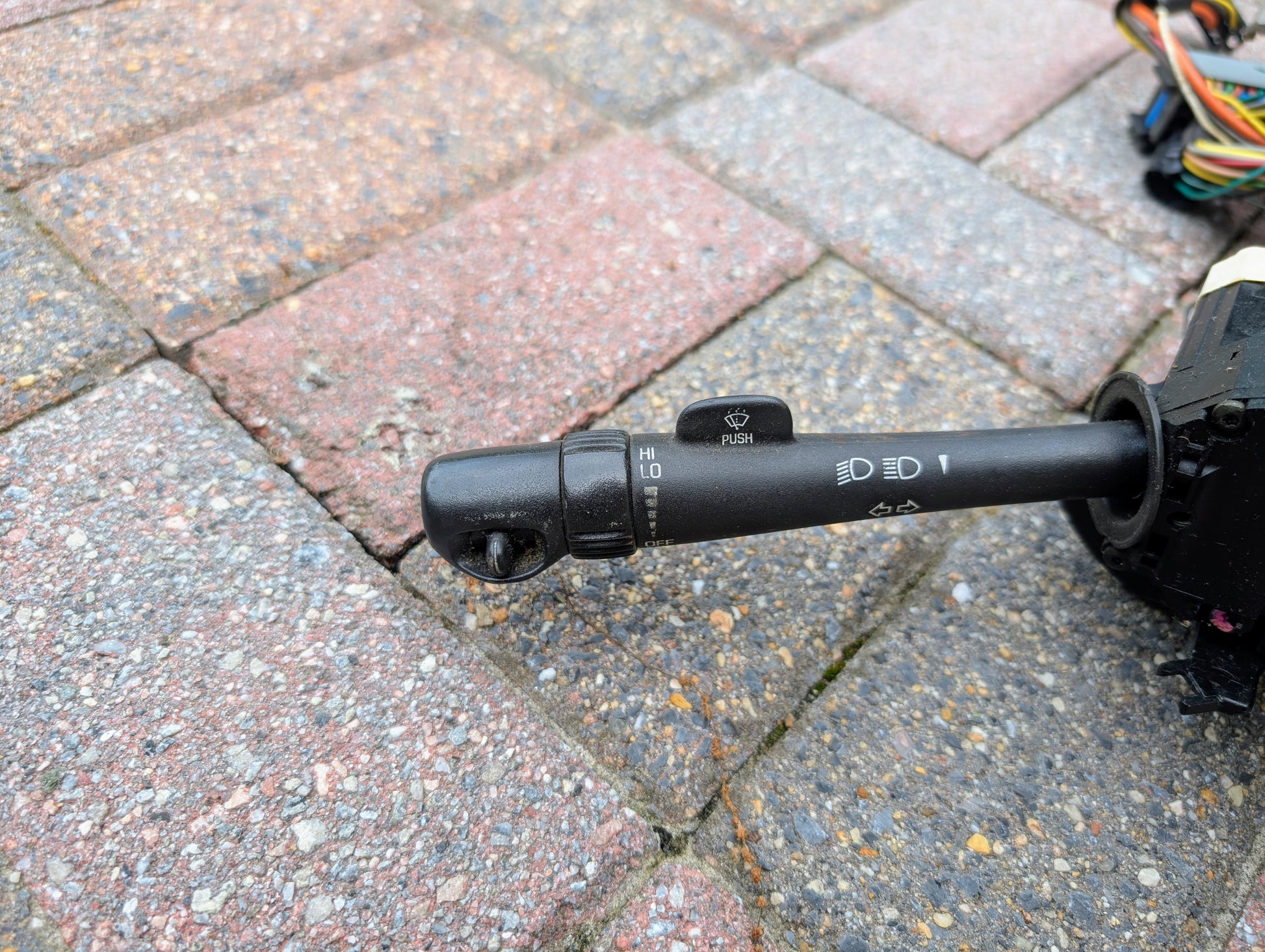 A close-up of a General Motors OEM Steering Column for 2000-2002 GM Full Size Trucks displays the multifunction turn signal and wiper lever on textured brick pavement, highlighting control symbols and some exposed wiring.