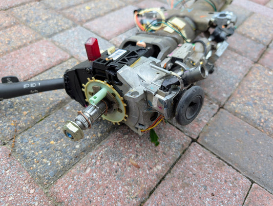 A close-up of a disassembled OEM Steering Column for 2000-2002 GM Full Size Trucks by General Motors, showing gears, wires, and electrical components on red and gray brick pavement.