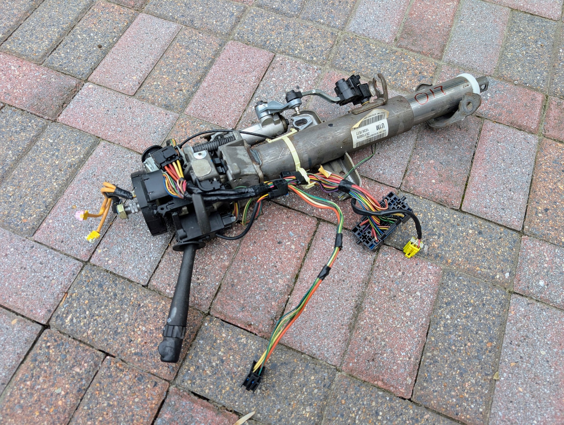 A disassembled General Motors OEM Steering Column for 2003-2006 GM Full Size Trucks, with wiring and connectors attached, is lying on a brick-paved surface.