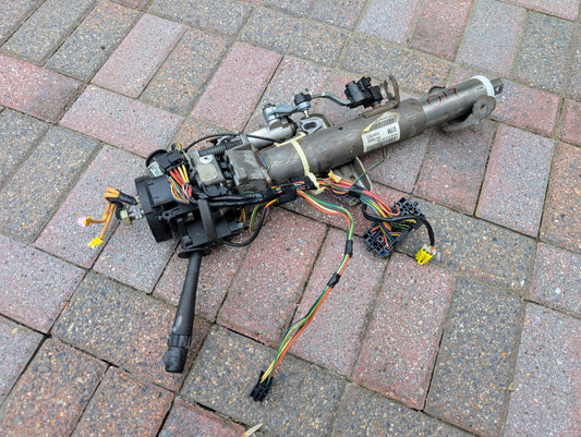 A disassembled General Motors OEM Steering Column for 2003-2006 GM Full Size Trucks, with wiring and connectors attached, is lying on a brick-paved surface.