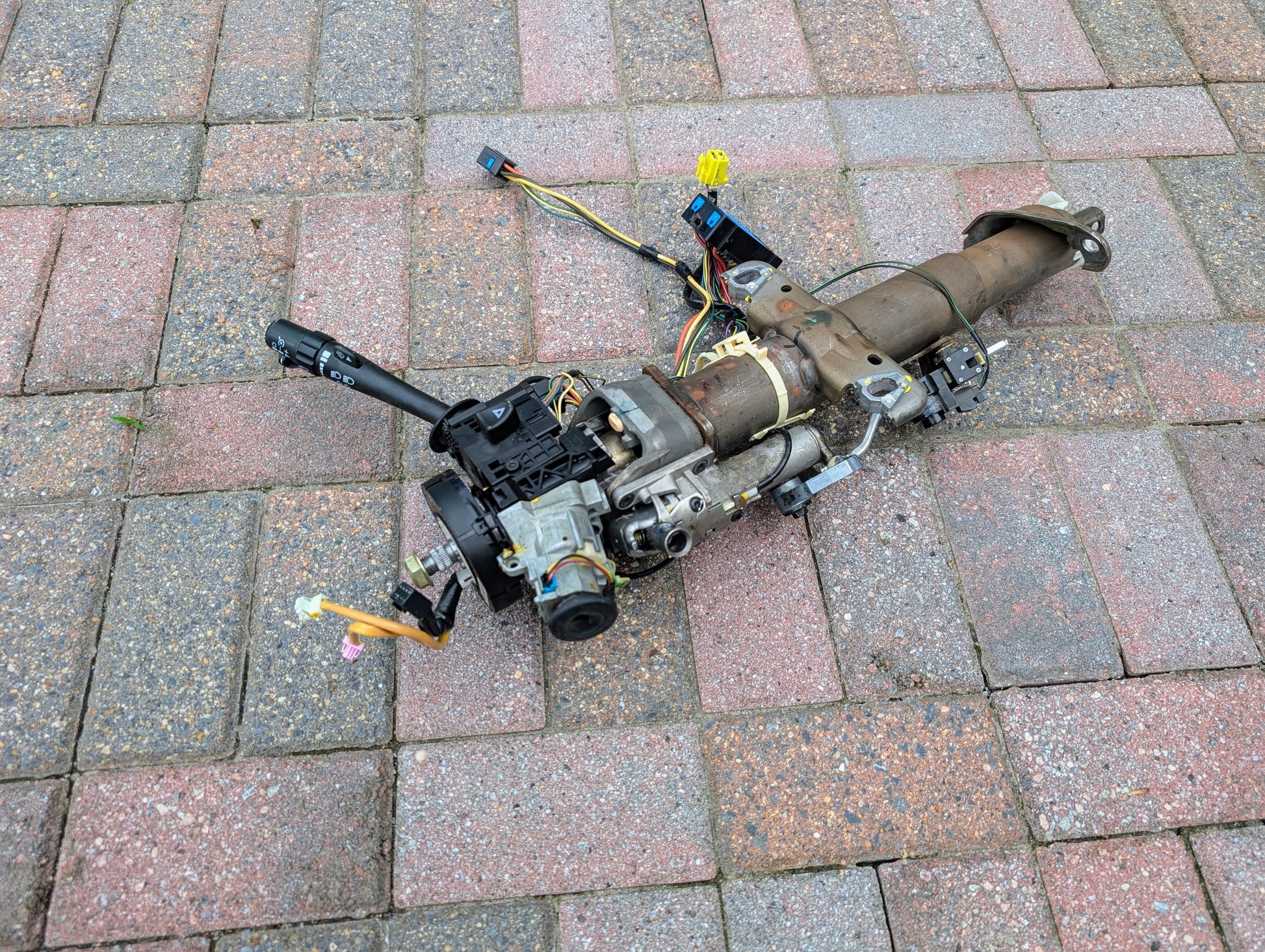 A General Motors OEM Steering Column for 2003-2006 GM Full Size Trucks, with wiring and controls for automatic transmission, is lying outdoors on a paved brick surface.