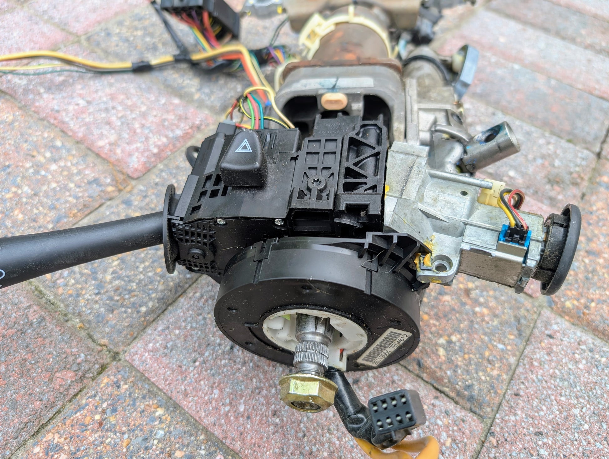A close-up of a General Motors OEM Steering Column for 2003-2006 GM Full Size Trucks with automatic transmission, displaying wiring, connectors, and mechanical parts on brick. The hazard light switch and turn signal stalk are visible.