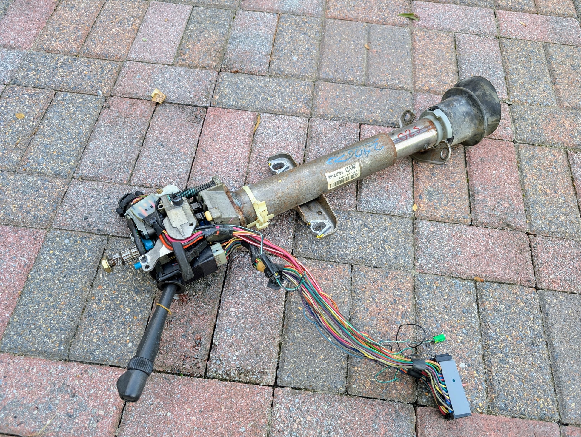 A General Motors OEM steering column with tilt, cruise control, and console shift for 1998-2005 Chevy S10 Blazer and more, with attached harness, rests on brick pavement after removal for repair or replacement.