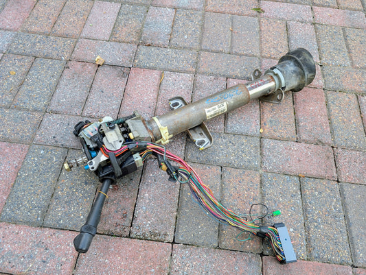 A General Motors OEM steering column with tilt, cruise control, and console shift for 1998-2005 Chevy S10 Blazer and more, with attached harness, rests on brick pavement after removal for repair or replacement.