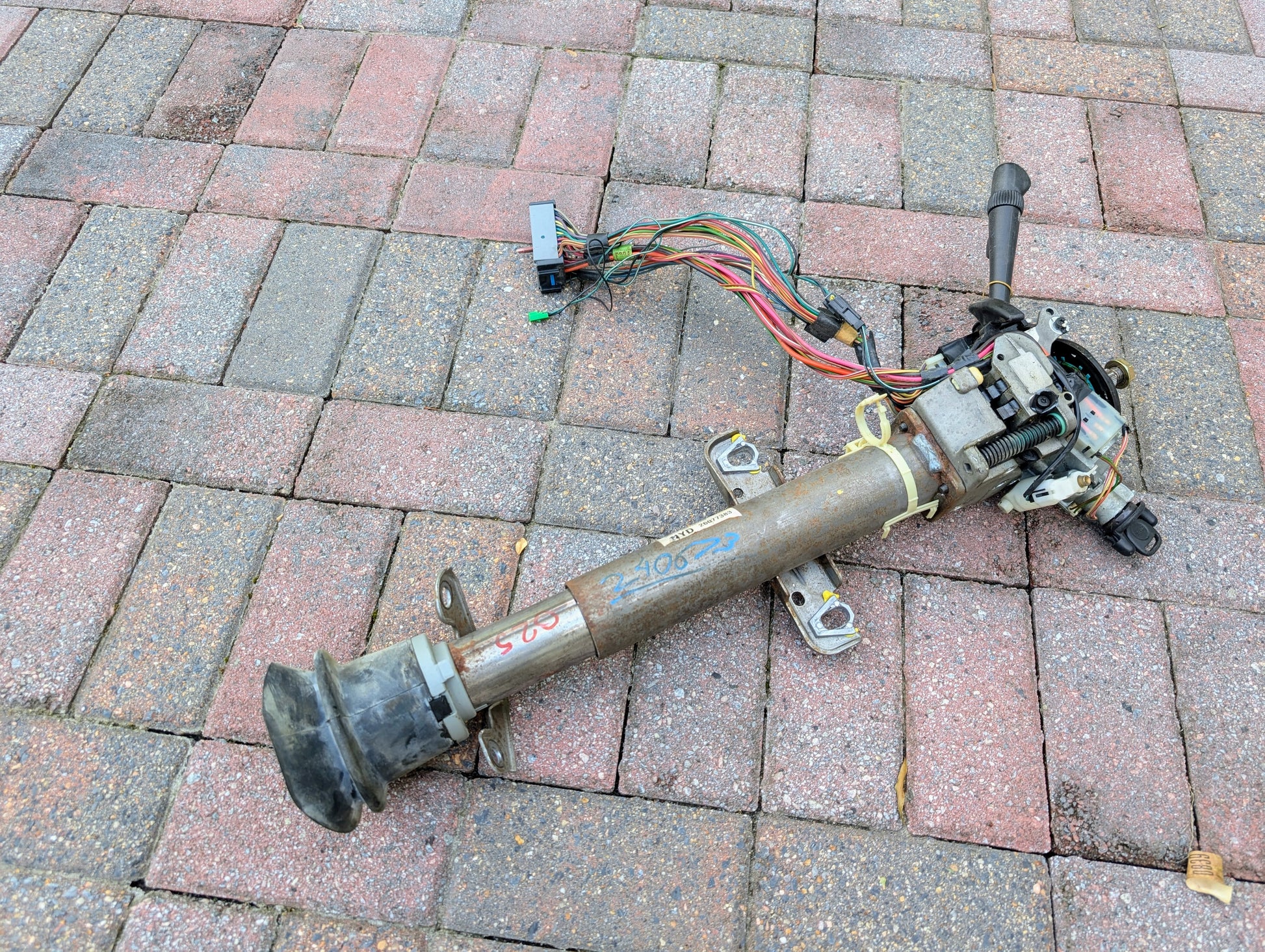 A used General Motors OEM Steering Column w/Tilt, Cruise Control, and Console Shift for 1998-2005 Chevy S10 Blazer lies on brick pavement. It has attached wires, a lever, colored writing, and visible signs of use.