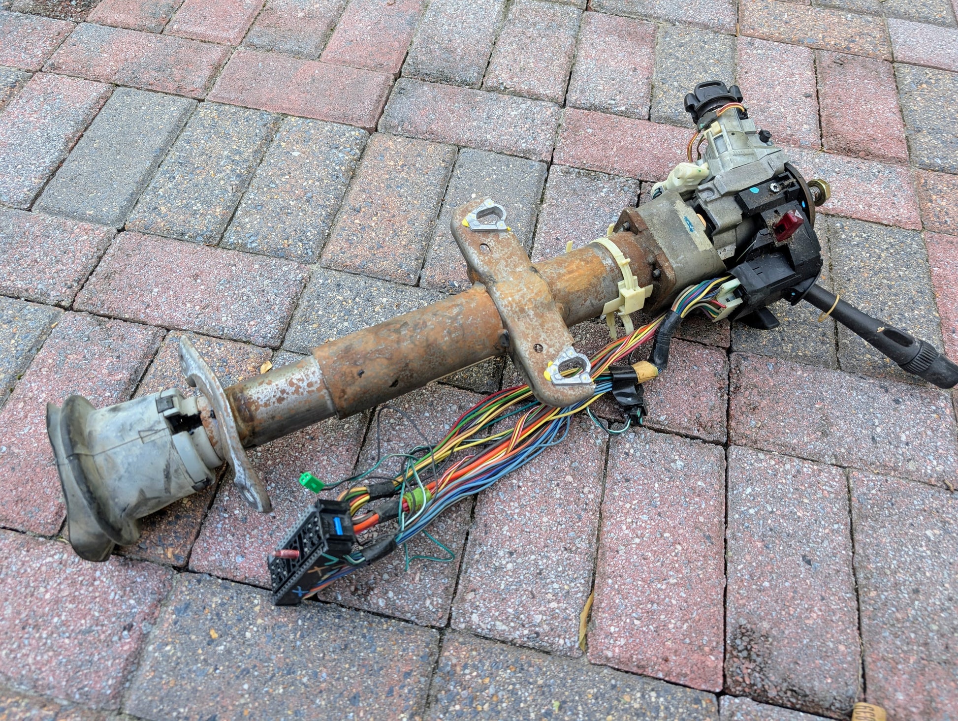 A General Motors OEM Steering Column with tilt, cruise control, and console shift for 1998-2005 Chevy S10 Blazer and more, complete with wiring and connectors, is shown resting on red and gray brick pavement.