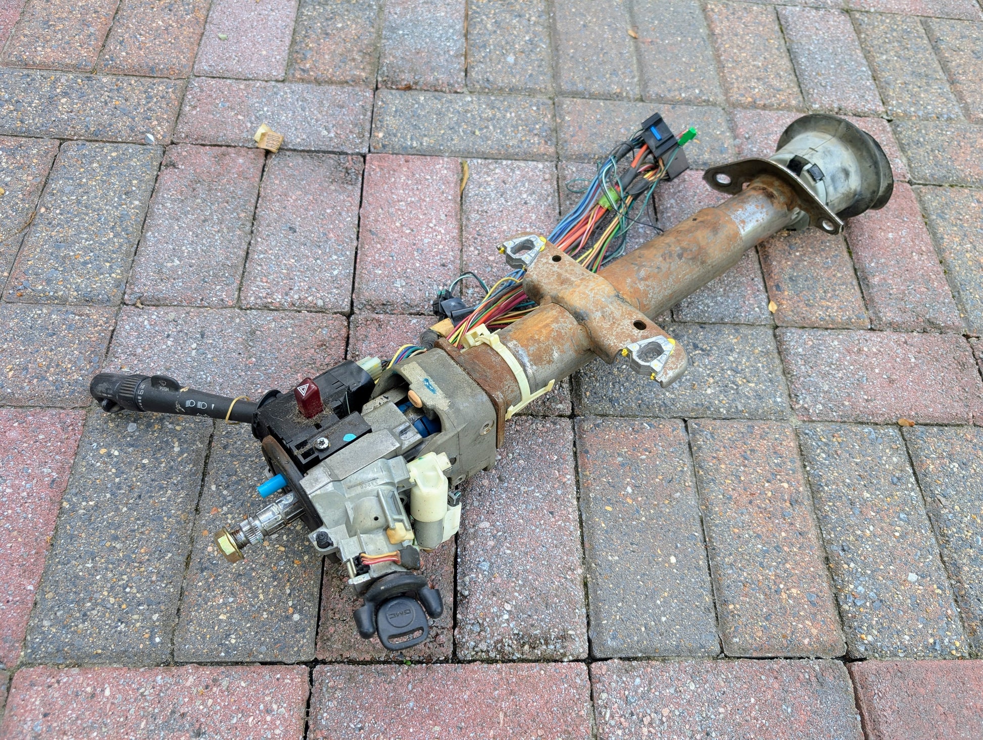 A General Motors OEM steering column with tilt, cruise control, and console shift for 1998-2005 Chevy S10 Blazer rests on a brick-paved surface.