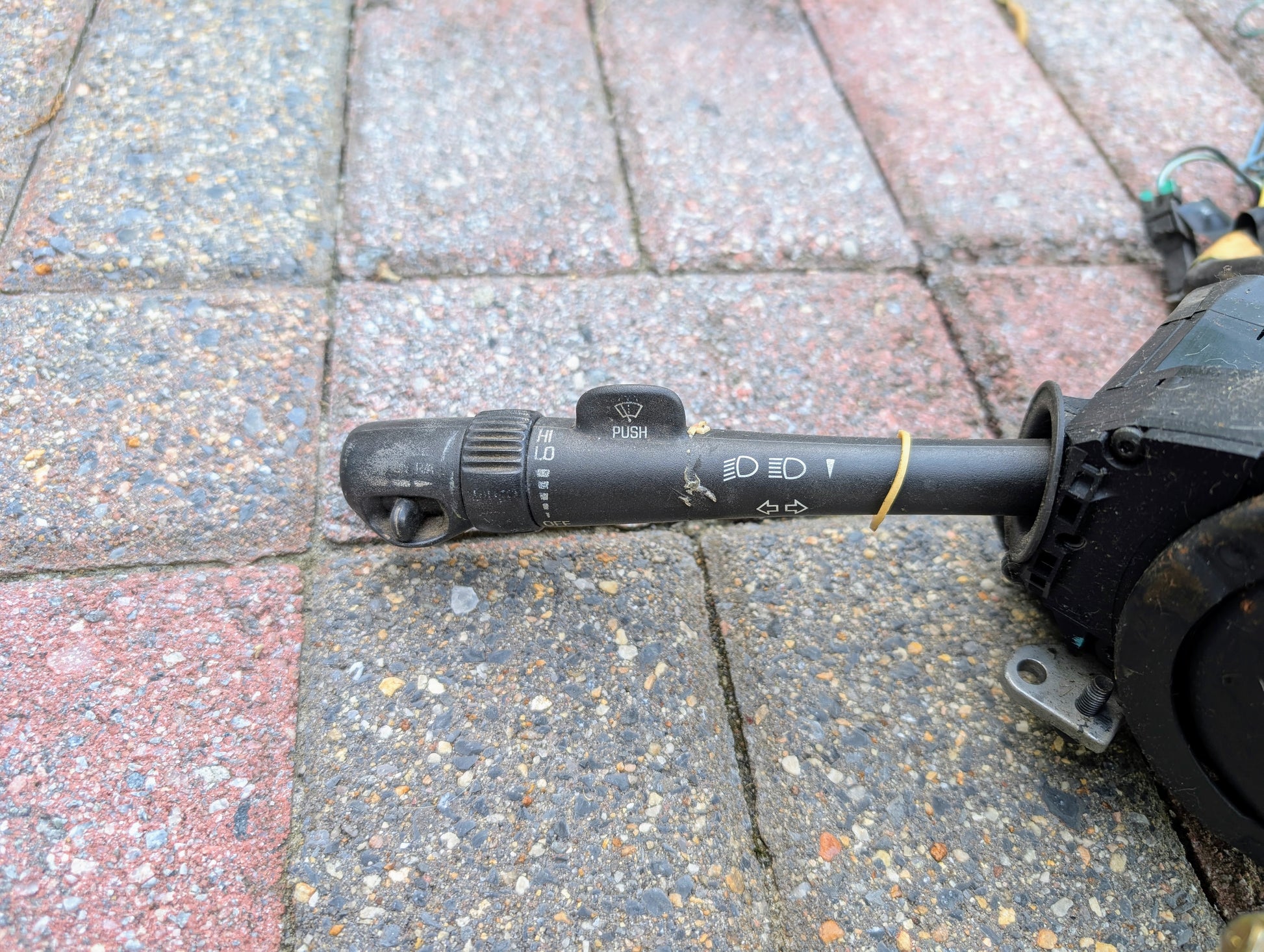 A used General Motors OEM Steering Column with tilt, cruise control, and console shift for 1998-2005 Chevy S10 Blazer and more lies on textured brick, showing slight wear. Black with white symbols and a PUSH button.