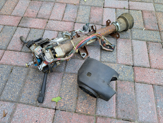 A used General Motors OEM Steering Column with Tilt, Cruise Control, and Column Shift for 1998-2005 Chevy S10 Blazer lies on pavement with attached wiring and a detached black plastic cover.