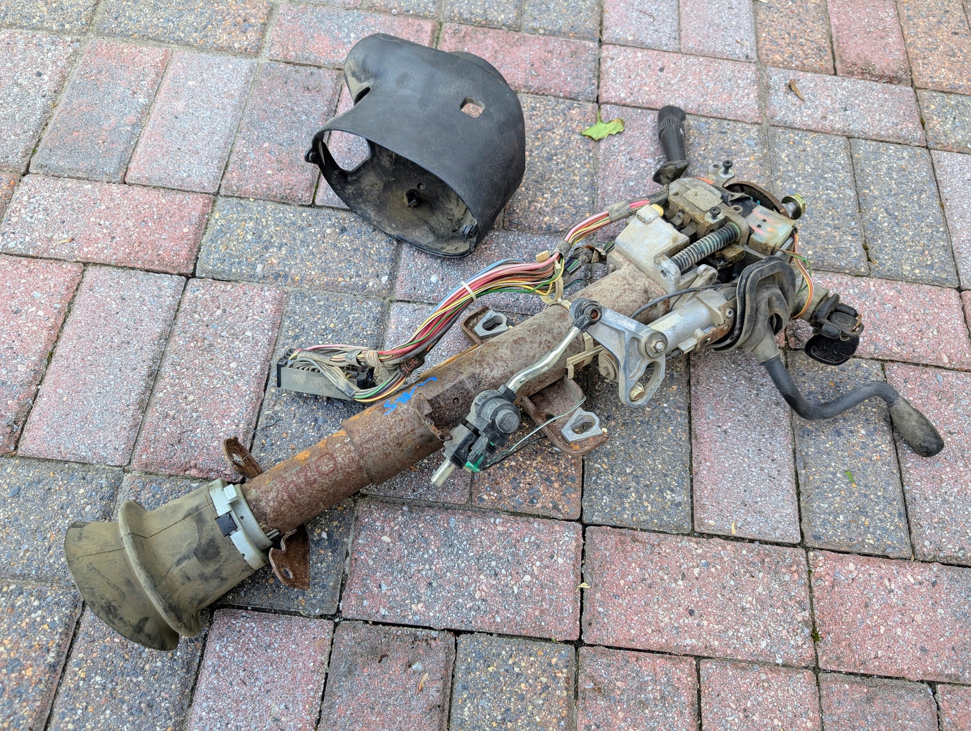 A General Motors OEM Steering Column with Tilt, Cruise Control, and Column Shift for 1998-2005 Chevy S10/Blazer lies disassembled with wiring and covers on a tiled outdoor surface.