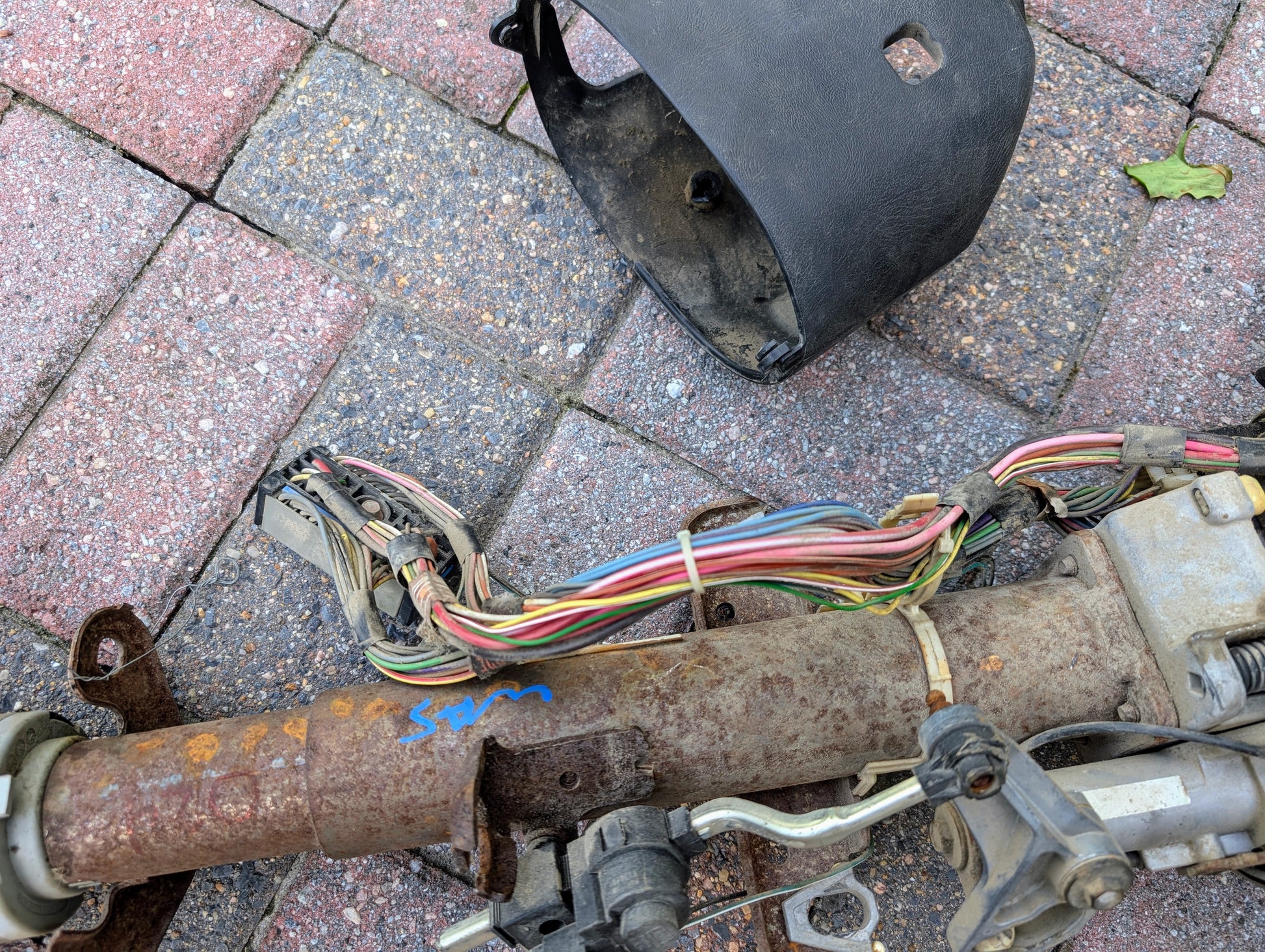 A close-up of a General Motors OEM Steering Column w/Tilt, Cruise Control, and Column Shift for 1998-2005 Chevy S10 Blazer, showing colorful wires and connectors outdoors on a brick pavement with the black cover detached.