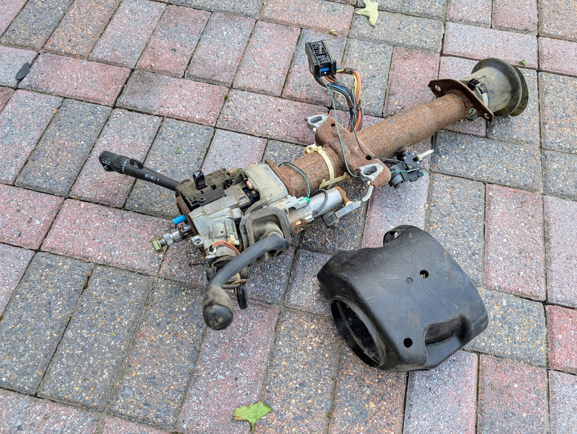 A General Motors OEM Steering Column w/Tilt, Cruise Control, and Column Shift for 1998-2005 Chevy S10/Blazer lies on pavement with attached levers, wiring, and a separate black plastic cover.