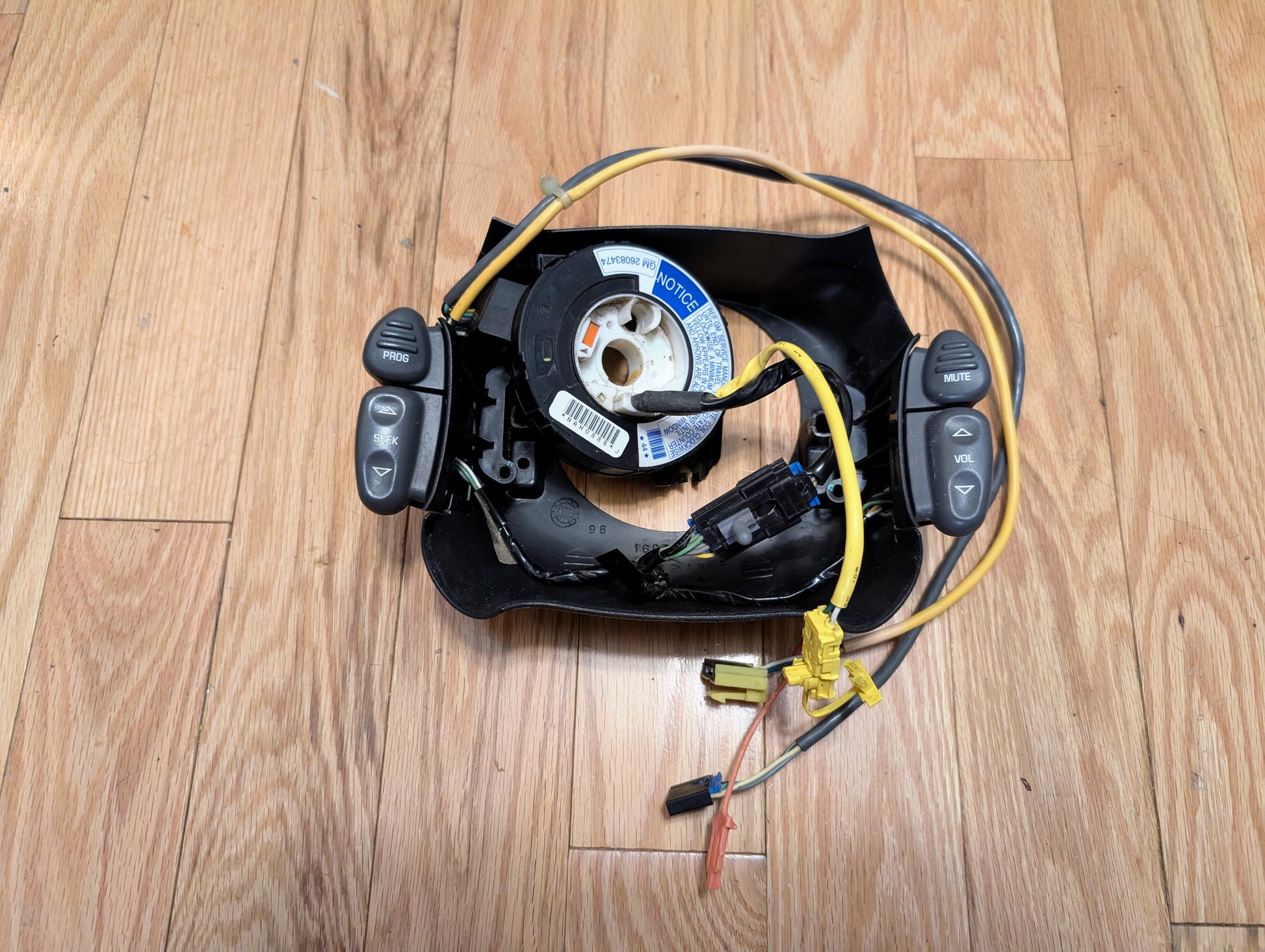 A disassembled General Motors KIT Steering Wheel Radio Controls & Clock Spring for 2000-2003 Chevy S10, Blazer, and more lies on a wooden floor with exposed wiring, radio buttons, cruise and volume controls visible.