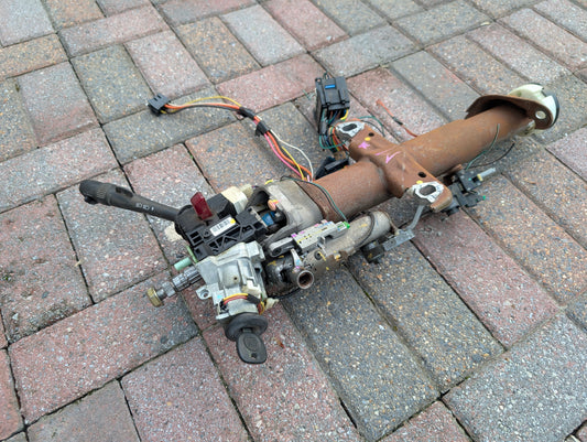 A General Motors OEM Steering Column w/KEY for 2000-2002 GM Full Size Trucks, with ignition switch, wiring, and automatic shifter attached, rests on red and gray brick pavement.