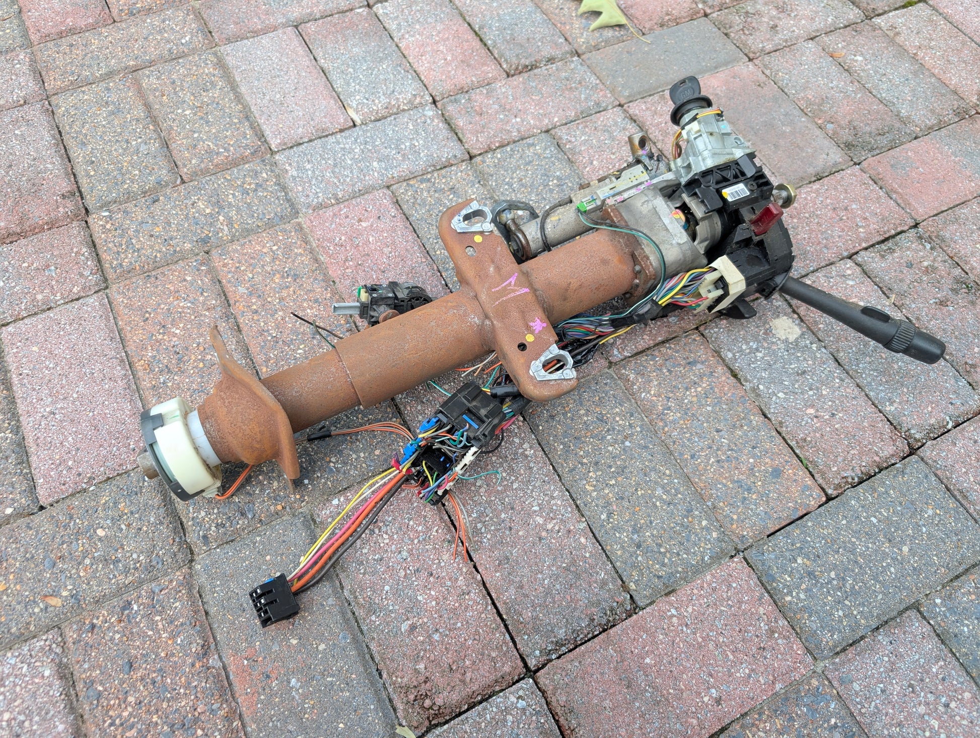 A rusty General Motors OEM Steering Column w/KEY for 2000-2002 GM Full Size Trucks, complete with wires, switches, and connectors, is lying on a brick-paved surface.