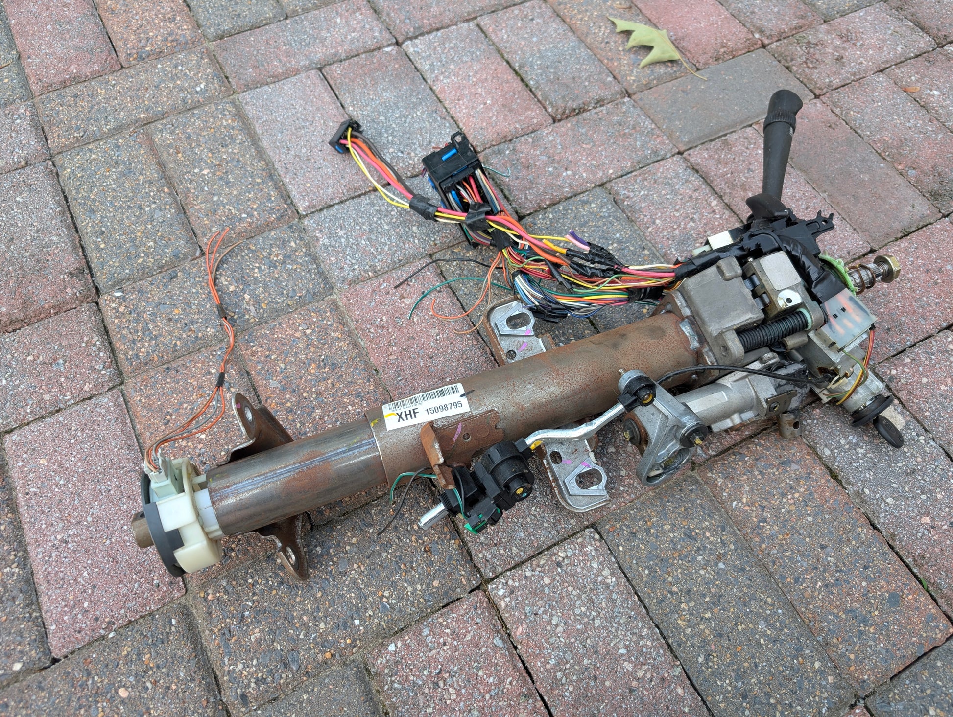 A General Motors OEM Steering Column w/KEY for 2000-2002 GM Full Size Trucks, featuring an automatic transmission column shifter, wiring, and connectors, is shown on a brick surface with visible shaft, mounting brackets, and wires.