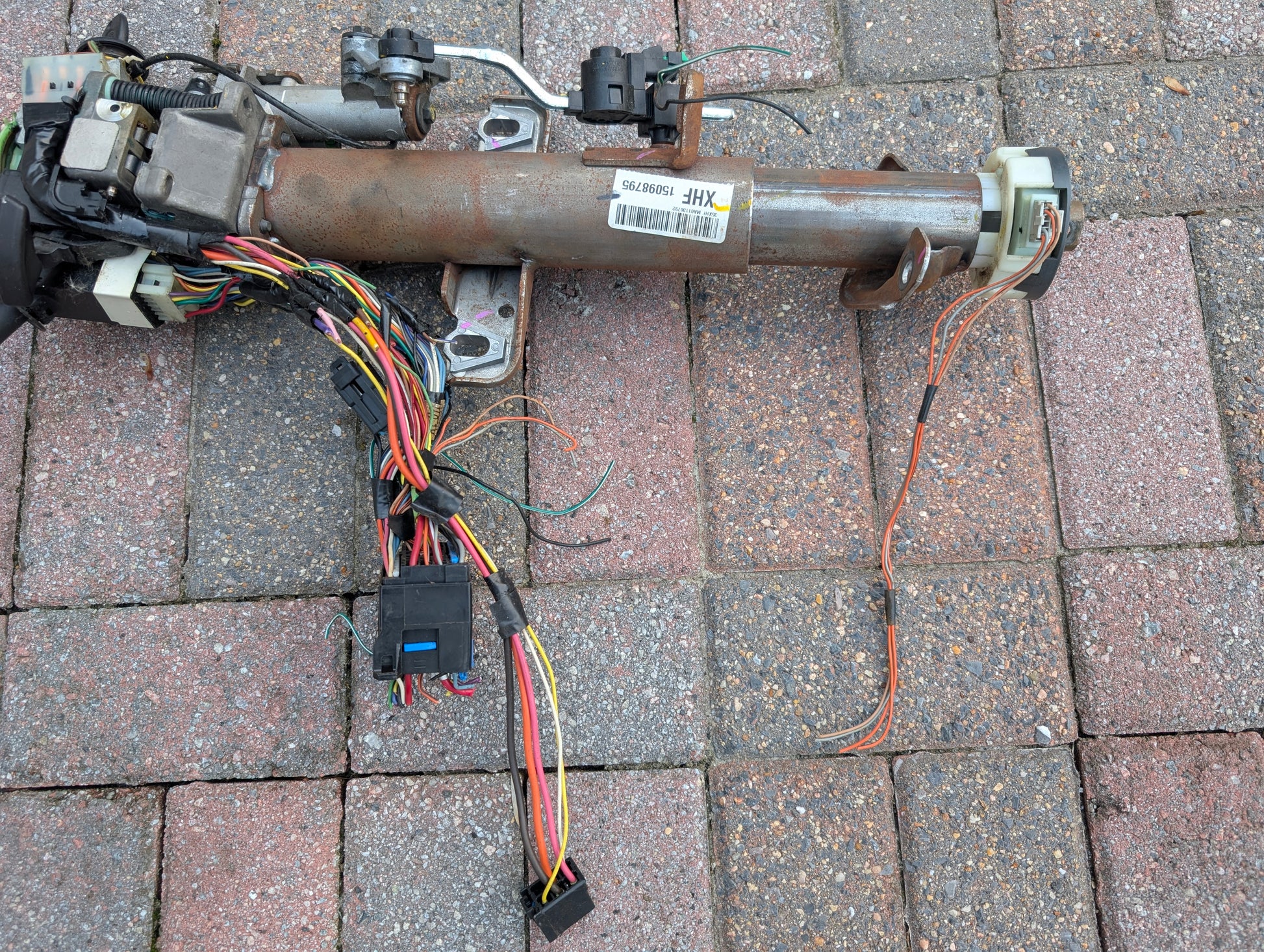 A General Motors OEM Steering Column w/KEY for 2000-2002 GM Full Size Trucks, featuring a wiring harness, connectors, and modules with visible colored wires, rests on a brick-paved ground.