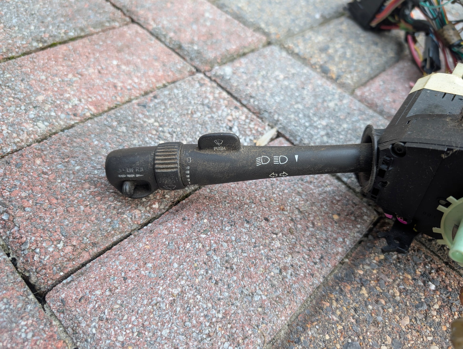A close-up of a General Motors OEM Steering Column w/KEY for 2000-2002 GM Full Size Trucks rests on a textured red and gray brick surface, with some car wiring and connectors partially visible in the background.