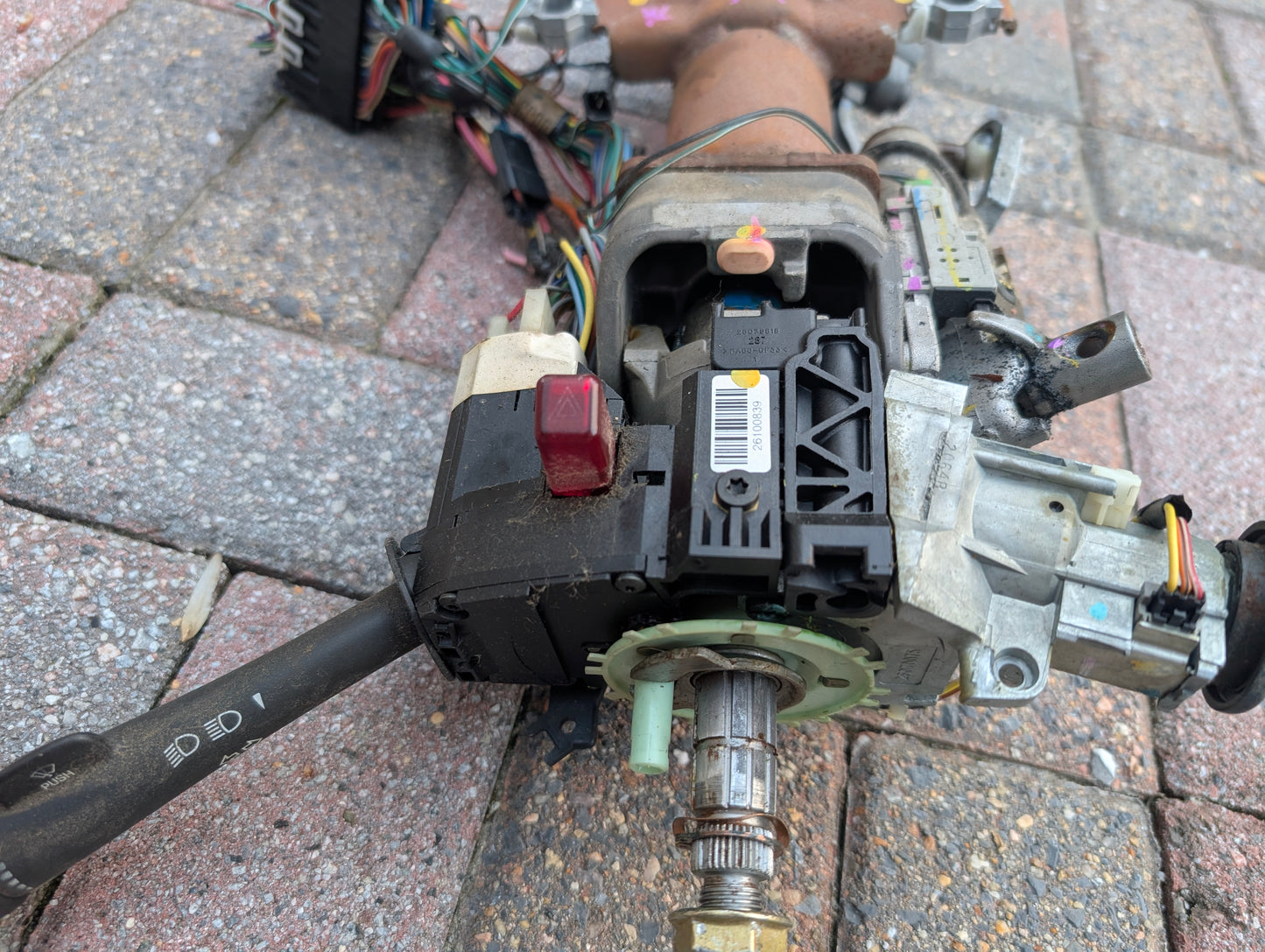 Close-up of a disassembled General Motors OEM Steering Column w/KEY for 2000-2002 GM Full Size Trucks, showing exposed wiring, connectors, ignition components, and column shifter, resting on brick pavement.