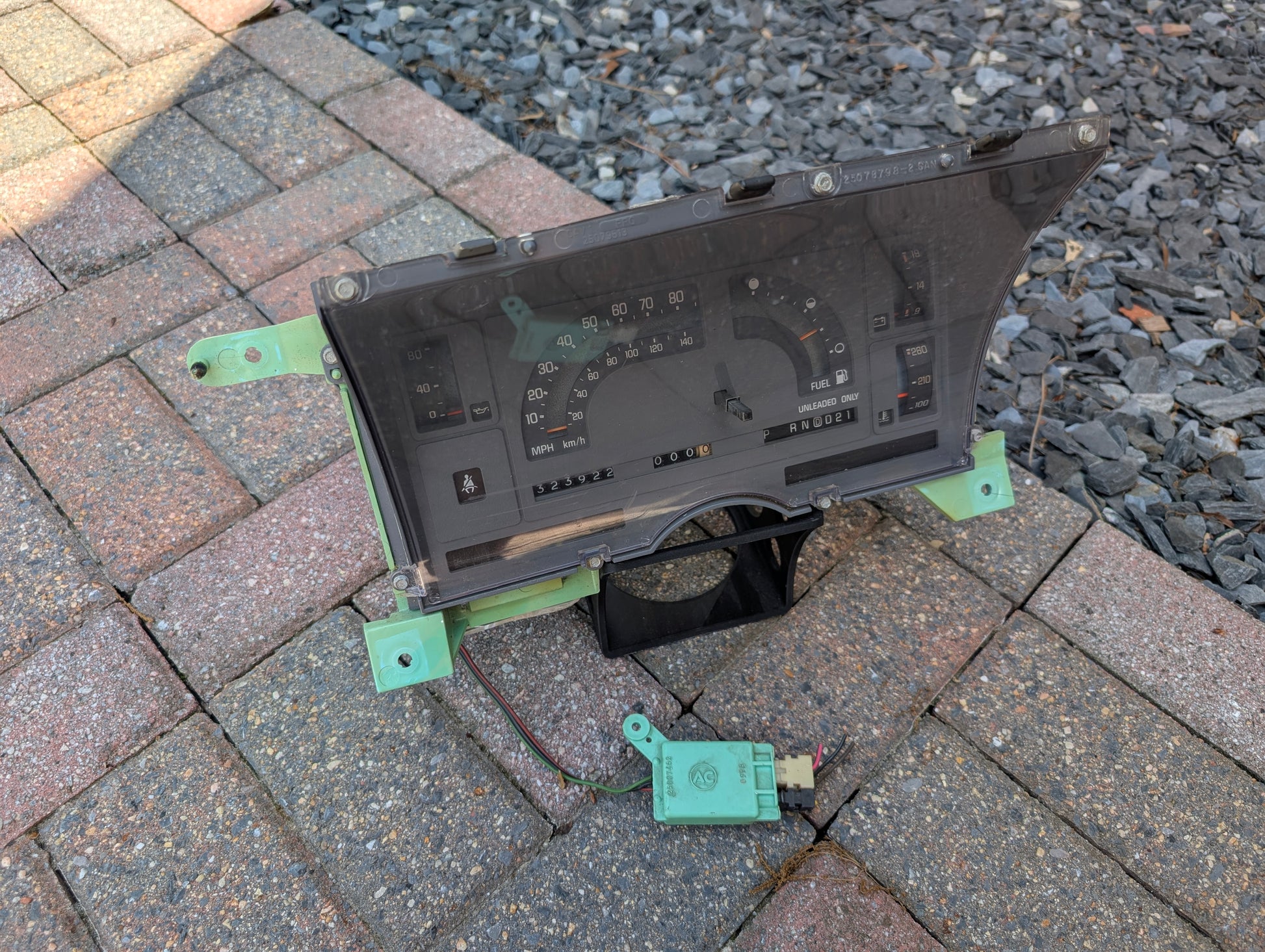 A General Motors OEM Instrument Cluster with Mechanical Speedo & PRNDL for 1986-1989 Chevy S10 Blazer and more is shown on a paved surface next to a small green speed sensor with wires; gravel appears in the background.