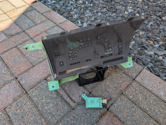 A General Motors OEM Instrument Cluster with Mechanical Speedo & PRNDL for 1986-1989 Chevy S10 Blazer and more is shown on a paved surface next to a small green speed sensor with wires; gravel appears in the background.