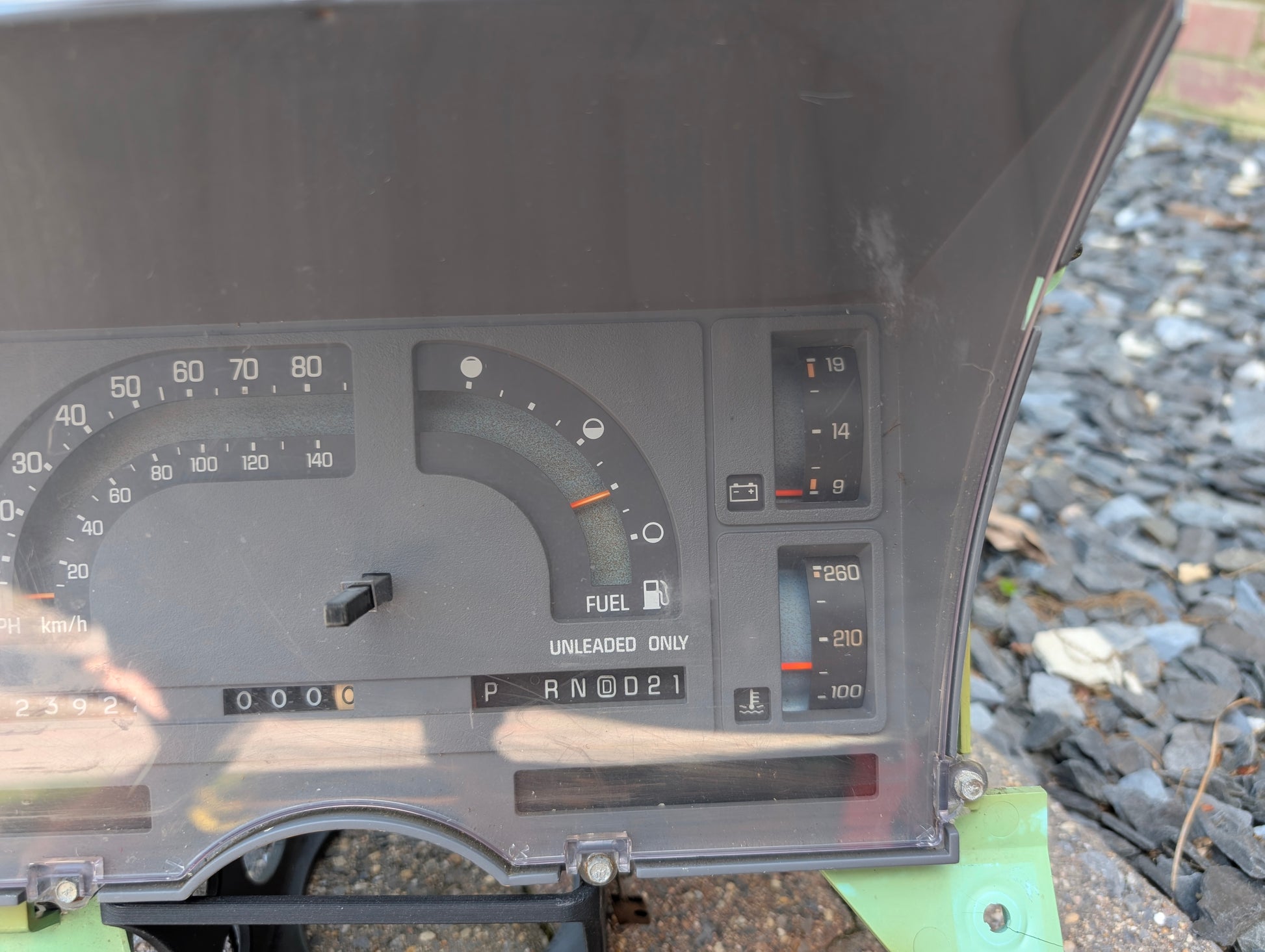 Close-up of the General Motors OEM Instrument Cluster with Mechanical Speedo & PRNDL for 1986-1989 Chevy S10 Blazer, showing speedometer, fuel and temperature gauges, gear indicator, and warning lights. Background includes gravel and a green vehicle panel.