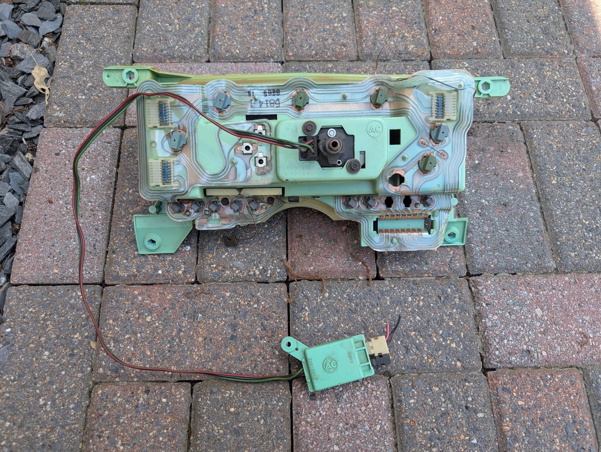 A green PCB with wiring and a small module, from a General Motors OEM Instrument Cluster with Mechanical Speedo & PRNDL (1986-1989 Chevy S10 Blazer), rests on brick pavement; it’s likely part of the vehicles dashboard assembly.