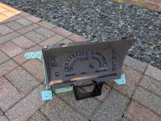 The General Motors Low Mile 52K OEM Instrument Cluster with PRNDL for 1986-1994 Chevy S10 Blazer and more is shown on brick pavement, featuring black and grey dials, a digital odometer, and gravel stones in the background.