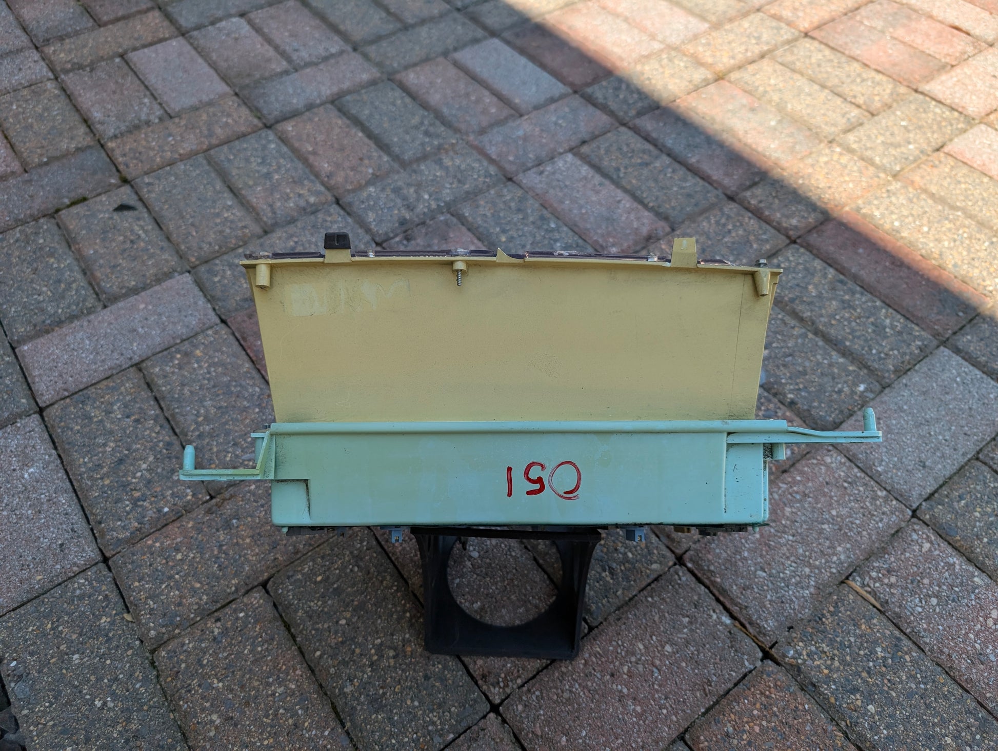 A weathered General Motors Low Mile 52K OEM Instrument Cluster with PRNDL for 1986-1994 Chevy S10 Blazer sits on a black stand outdoors on brick pavement, featuring light green and yellow details and ISQ in red.
