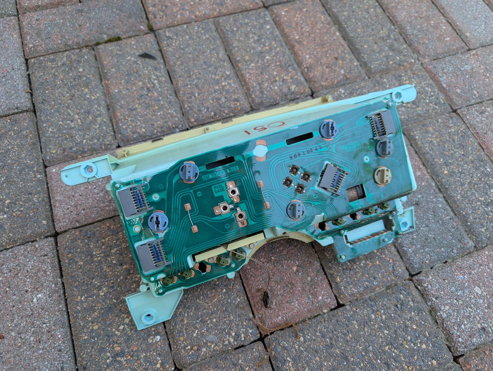 A disassembled General Motors 52K OEM Instrument Cluster with PRNDL for 1986-1994 Chevy S10 Blazer and more rests on rectangular brick pavement, showing its exposed circuit board.
