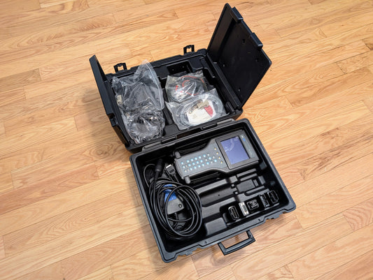 A black plastic case on a wooden floor holds a Vetronix TESTED Tech 2 Automotive Diagnostic Tool & Accessories for 1991–2013 GM vehicles, with keypad, screen, cables, adapters, and accessories organized in compartments.