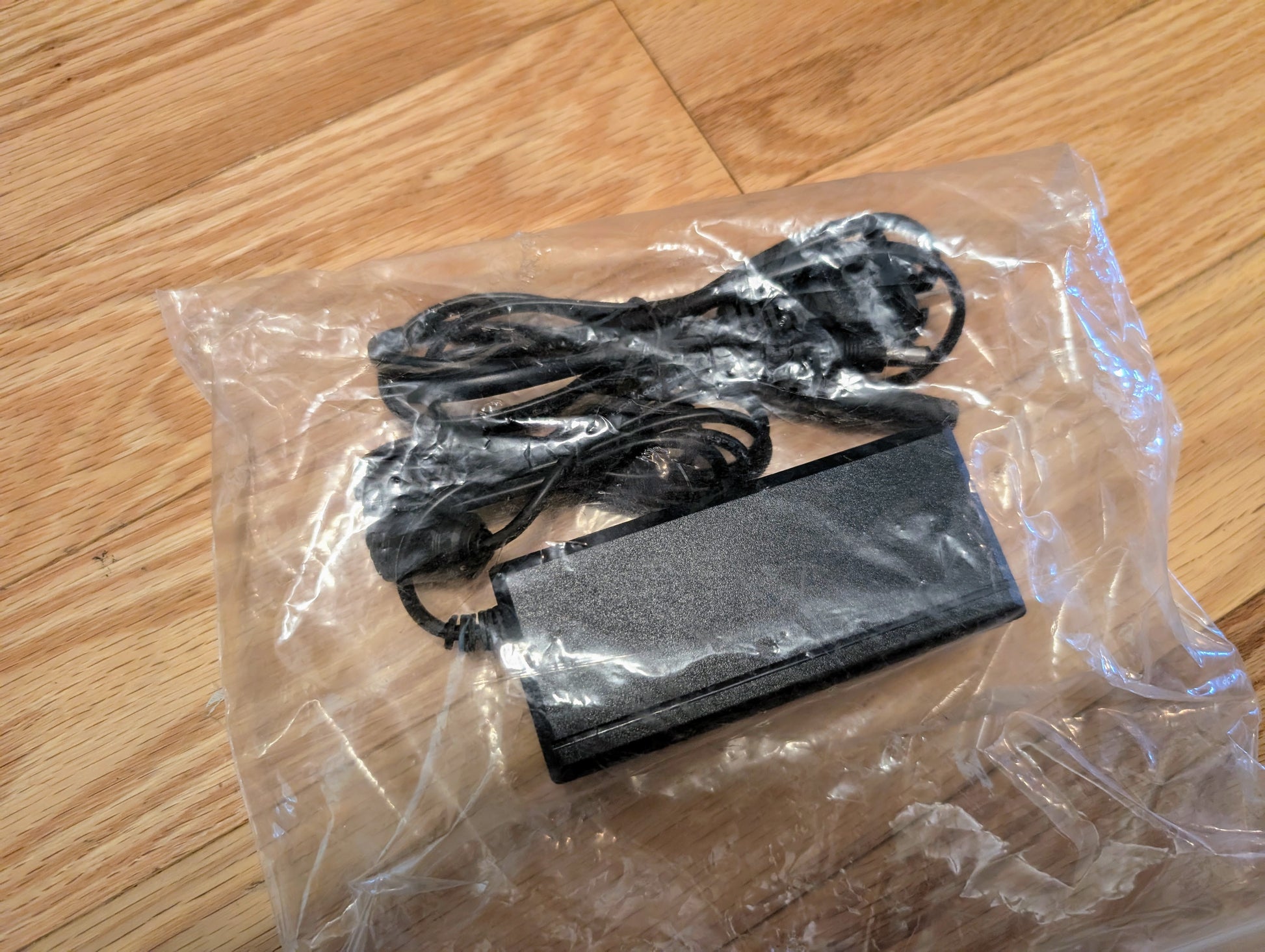 A black power adapter with a coiled cable, packaged in clear plastic, sits on a wooden floor—ideal for powering your Vetronix TESTED Tech 2 Automotive Diagnostic Tool & Accessories for 1991-2013 GM vehicles.