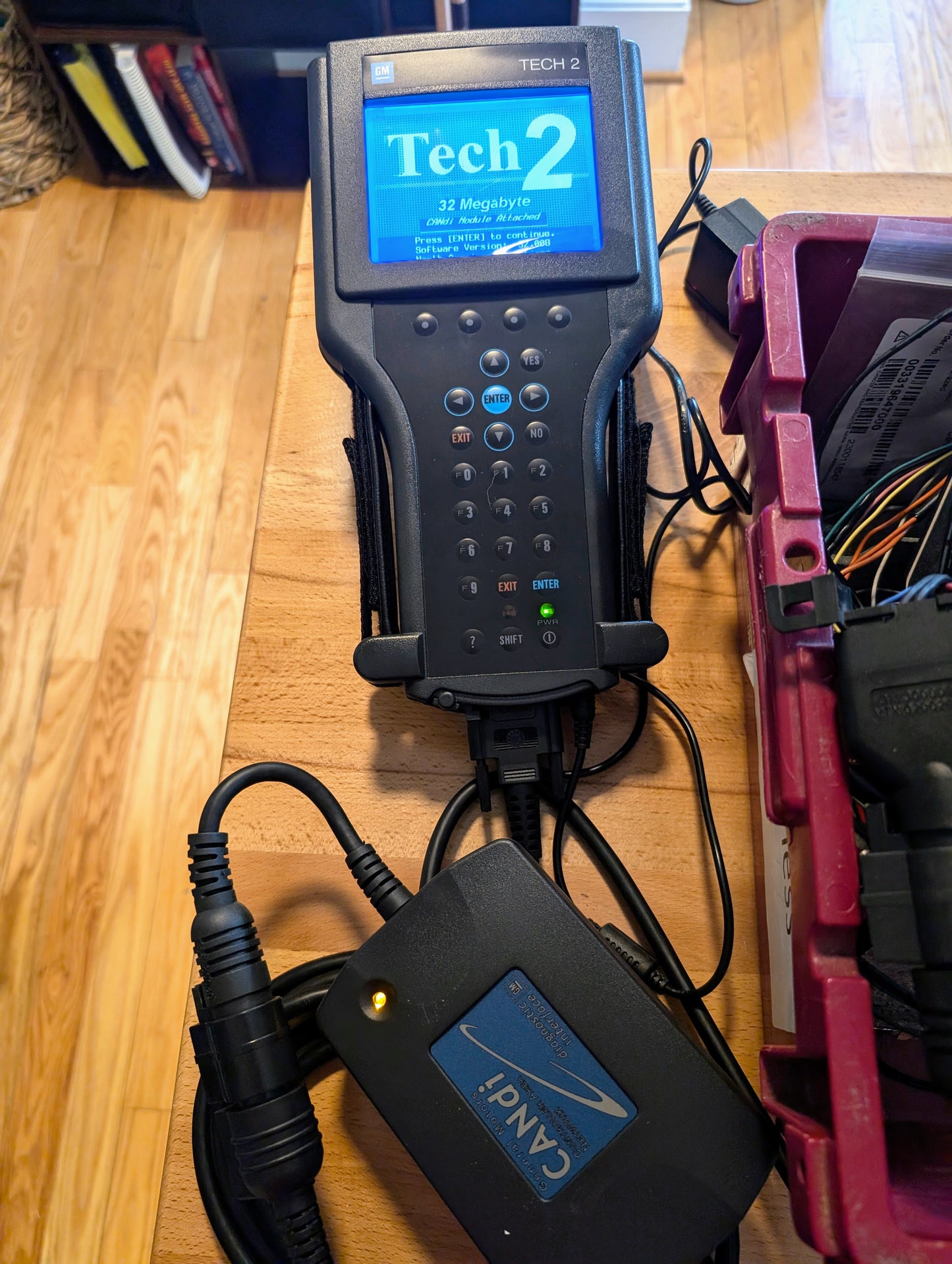A Vetronix TESTED Tech 2 Automotive Diagnostic Tool & Accessories for 1991-2013 GM vehicles, featuring a blue display and attached cables, rests on a wooden table beside a red plastic toolbox.