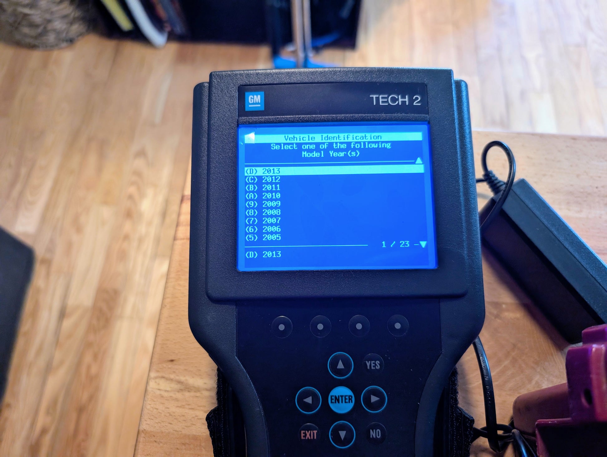 The Vetronix TESTED Tech 2 Automotive Diagnostic Tool & Accessories for 1991-2013 GM vehicles, an OBD2 code scanner, displays a blue screen with selectable model years and sits on a wooden surface, ready for professional use.