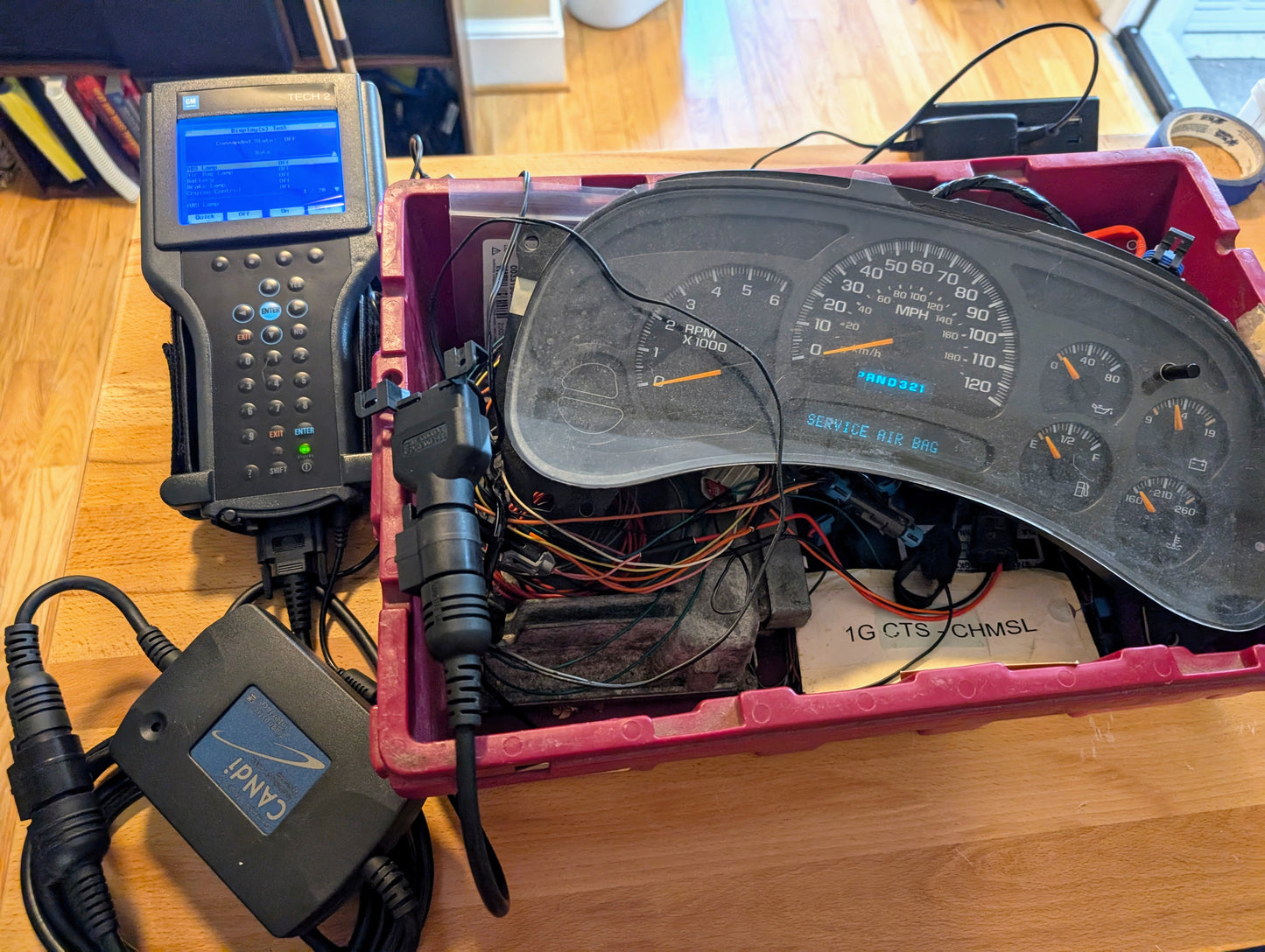 A Vetronix TESTED Tech 2 Automotive Diagnostic Tool & Accessories for 1991-2013 GM Vehicles is connected to a vehicles instrument cluster with exposed wiring, placed in a red plastic bin; a handheld OBD2 code scanner with blue screen is attached.