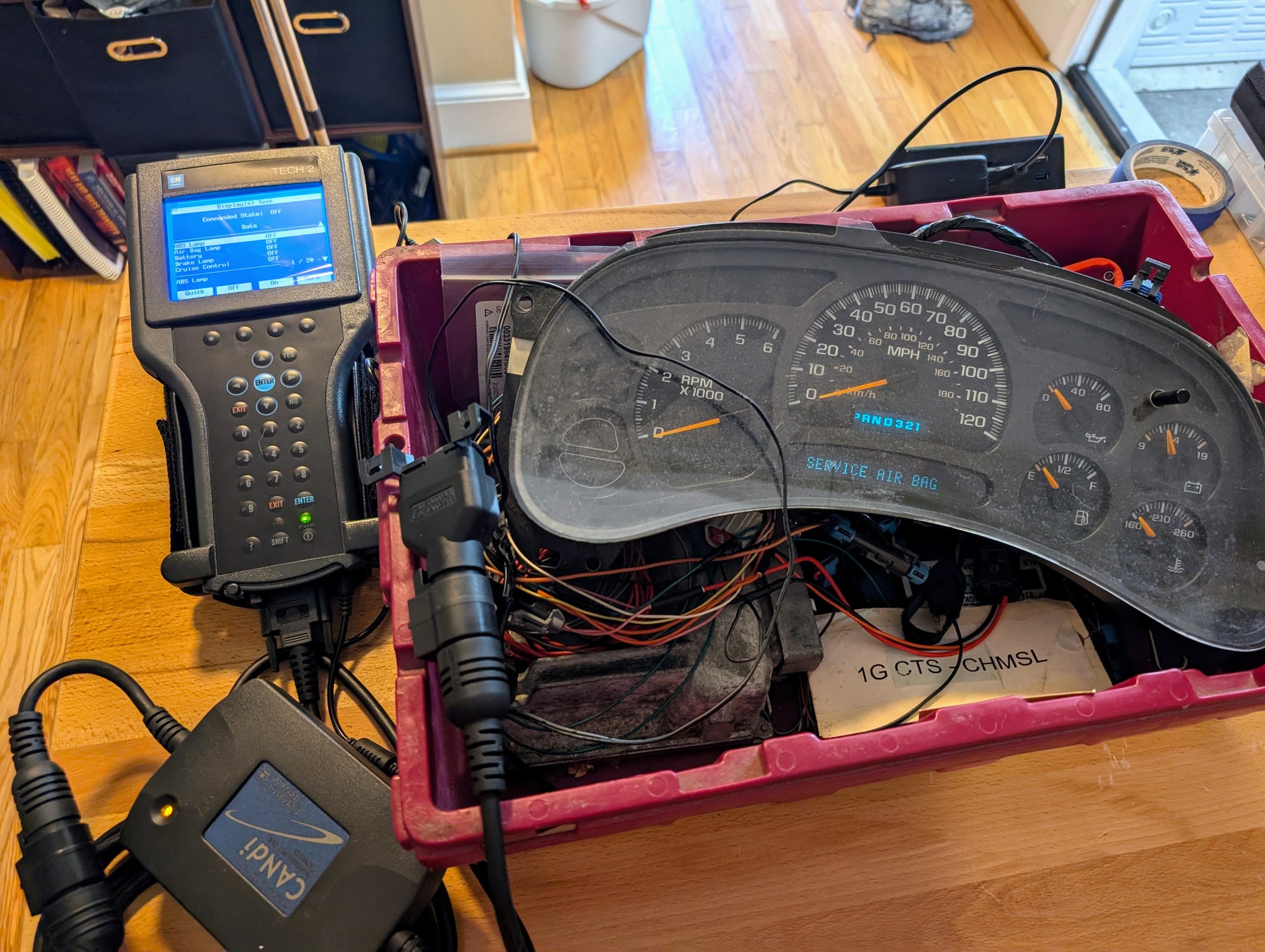 A Vetronix TESTED Tech 2 Automotive Diagnostic Tool & Accessories for 1991-2013 GM Vehicles displays data while connected to a vehicle instrument cluster and wiring, all inside a pink container on a wooden table.