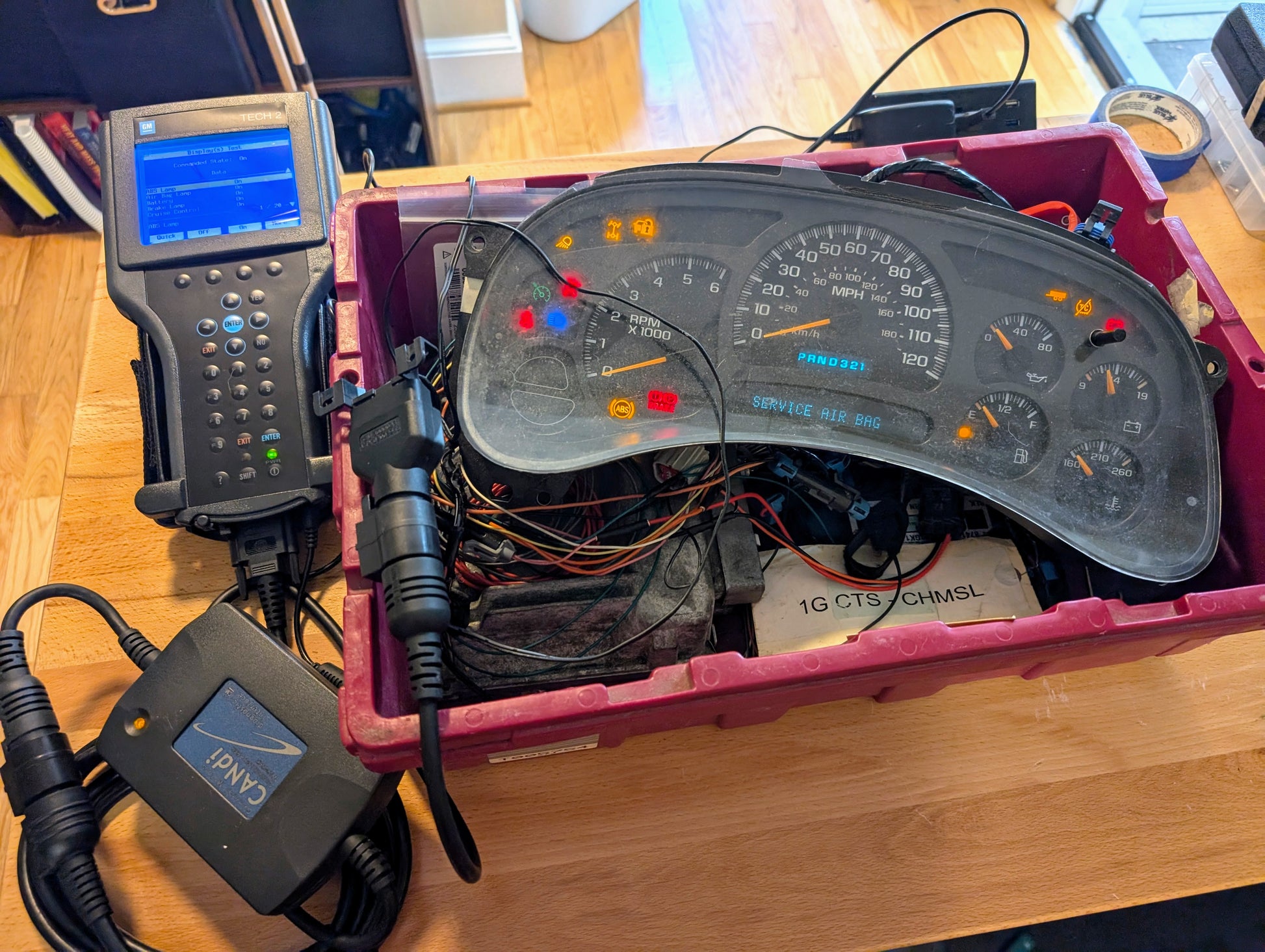 A Vetronix TESTED Tech 2 Automotive Diagnostic Tool & Accessories for 1991-2013 GM Vehicles are connected to a car dashboard instrument cluster, all set in a red plastic bin on a wooden floor with various diagnostic cables and equipment nearby.
