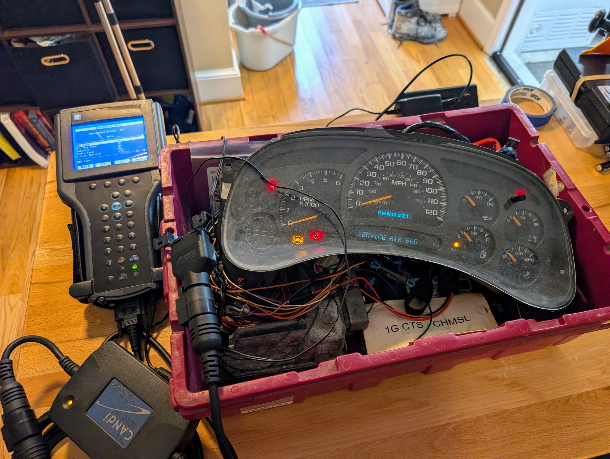 A Vetronix TESTED Tech 2 Automotive Diagnostic Tool & Accessories for 1991-2013 GM Vehicles, connected to a car dashboard instrument cluster via cables, sits on a wooden table alongside electronic equipment and a handheld OBD2 code scanner. Household items are in the background.