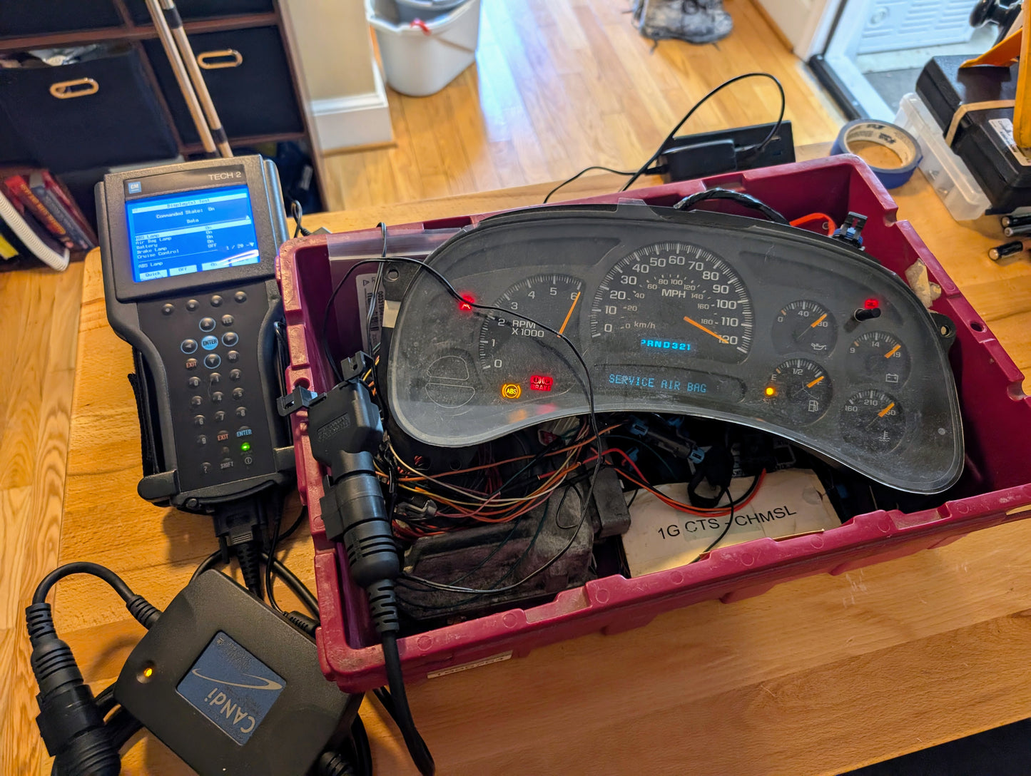 A Vetronix TESTED Tech 2 Automotive Diagnostic Tool & Accessories for 1991-2013 GM vehicles connects to a car dashboard instrument cluster on a wooden table, with wires and devices visible, indicating testing or repair.