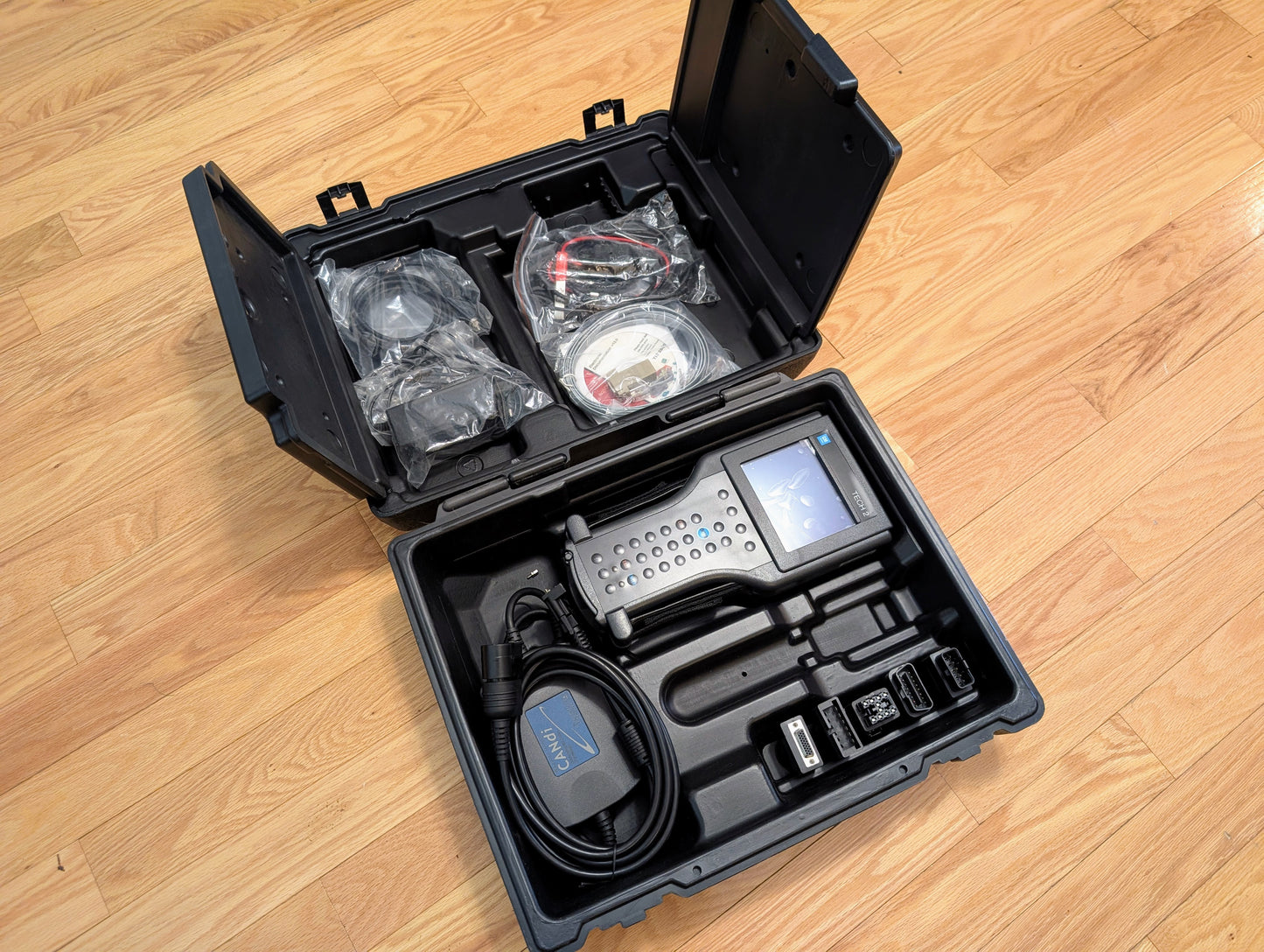 A rugged black case on a wooden floor holds a Vetronix TESTED Tech 2 Automotive Diagnostic Tool & Accessories for 1991-2013 GM Vehicles, including screen, cables, connectors, and accessories arranged in molded compartments.
