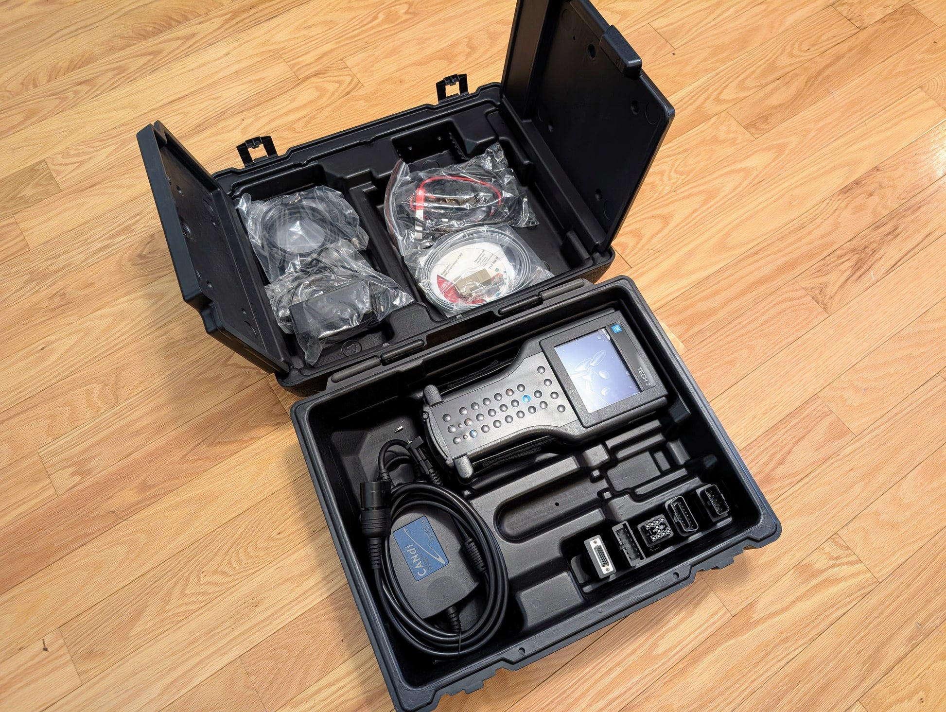 A rugged black case on a wooden floor holds a Vetronix TESTED Tech 2 Automotive Diagnostic Tool & Accessories for 1991-2013 GM Vehicles, including screen, cables, connectors, and accessories arranged in molded compartments.