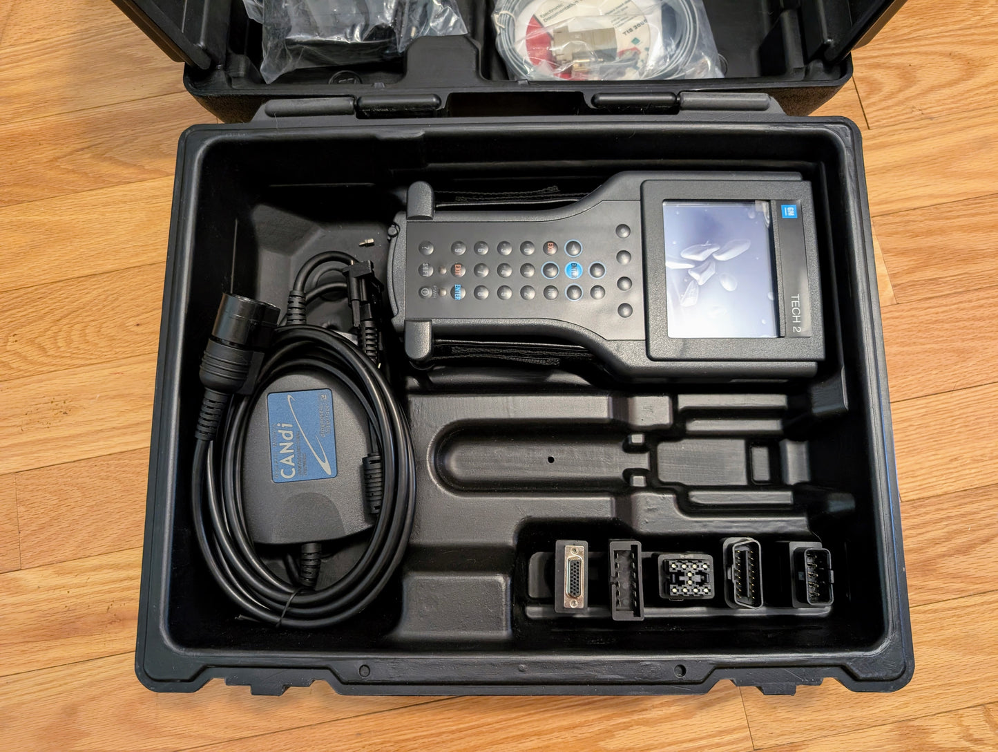 A black plastic case holds a Vetronix TESTED Tech 2 Automotive Diagnostic Tool & Accessories for 1991-2013 GM vehicles, including cables, a CANDi OBD2 adapter, and connectors, all arranged in foam slots on a wooden floor.