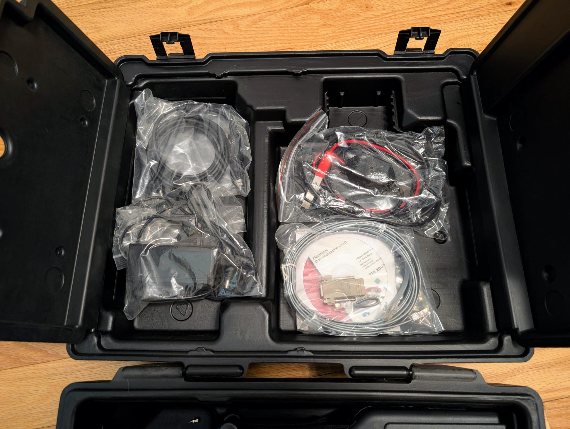 A black plastic case with compartments holds Vetronix TESTED Tech 2 Automotive Diagnostic Tool & Accessories for 1991-2013 GM Vehicles, including cables, adapters, and discs—all sealed in clear bags on a wooden surface.