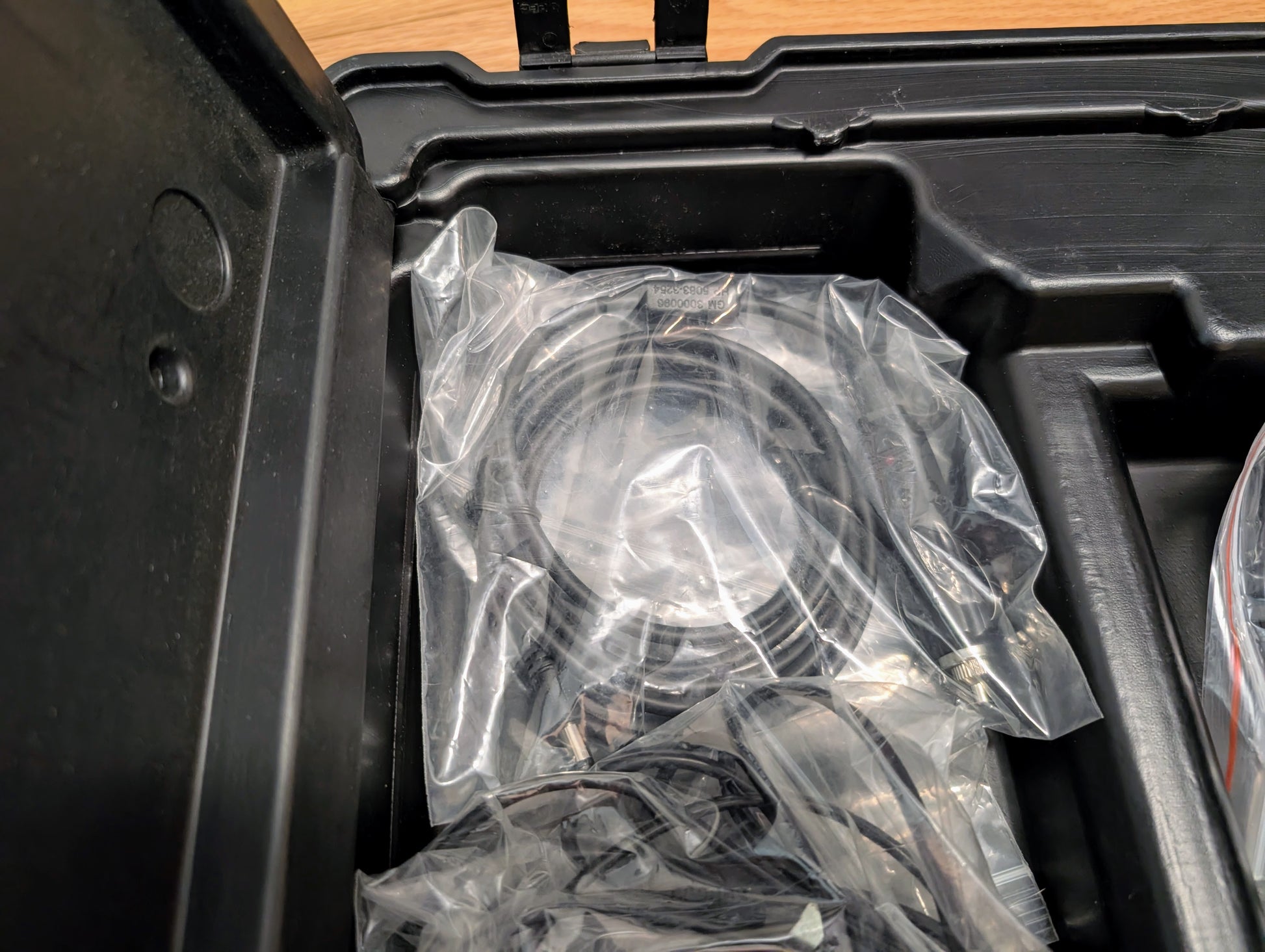 A round object in a clear plastic bag is placed inside a black hard case with molded compartments, revealing storage for the Vetronix TESTED Tech 2 Automotive Diagnostic Tool & Accessories for 1991-2013 GM Vehicles.
