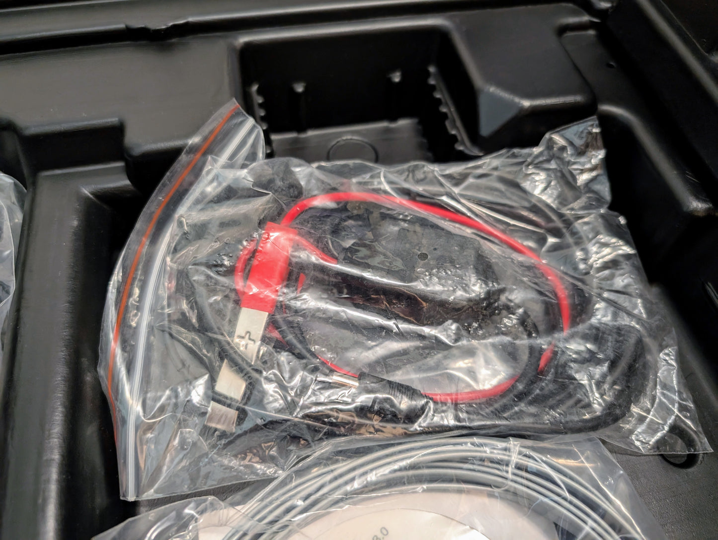 A red and black USB cable is sealed in a clear plastic bag and placed in a black tray with other items included with the Vetronix TESTED Tech 2 Automotive Diagnostic Tool & Accessories for 1991-2013 GM vehicles.