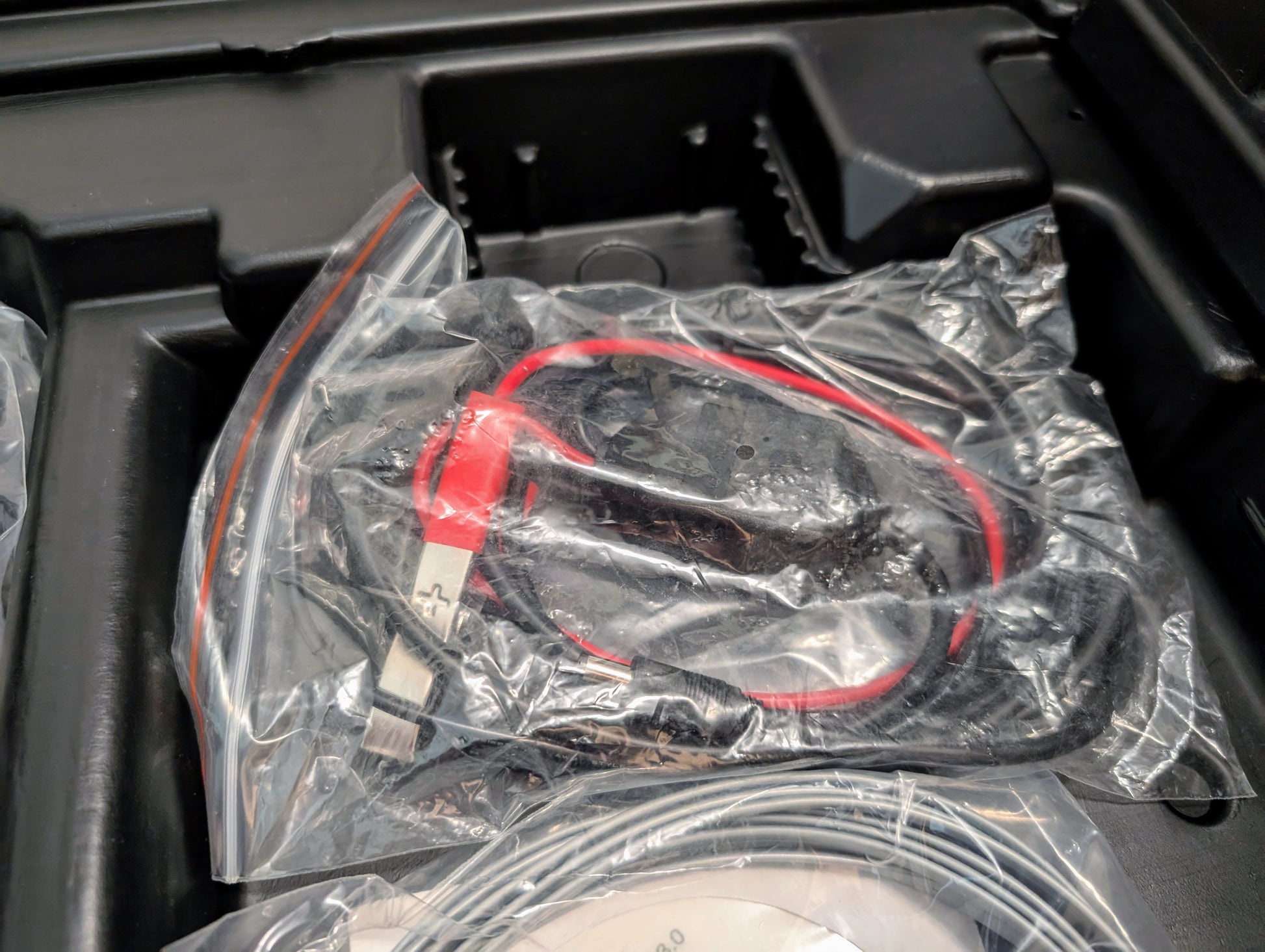 A red and black USB cable is sealed in a clear plastic bag and placed in a black tray with other items included with the Vetronix TESTED Tech 2 Automotive Diagnostic Tool & Accessories for 1991-2013 GM vehicles.